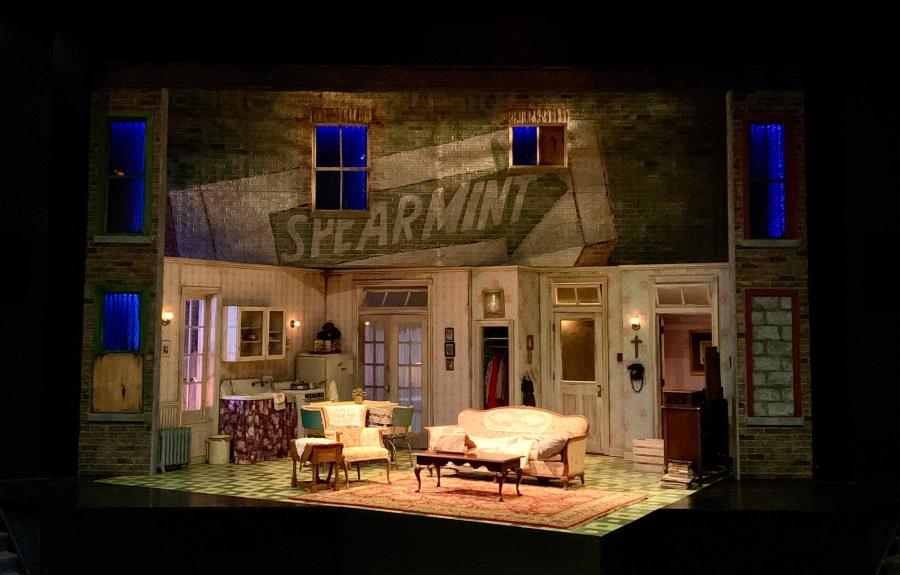 A Raisin in the Sun scenic design on stage, including a room filled with mid 20th century furniture: an armchair and matching couch, two coffee tables, a sink, stove, refrigerator, closet, radiator, a cross on the wall above a telephone, a stack of books on the floor, and a brick wall above the set with a painted “Spearmint” ad