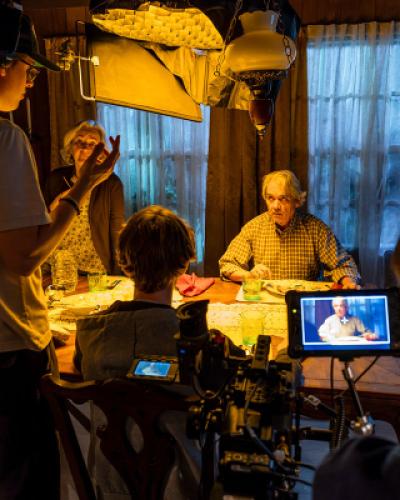 In foreground, a student filmmaker wearing headphones talks to actors during the filming of the movie Remembering Colin Stall. In background over a dining table set, three actors are being filmed having a meal. On right, a film camera shows a frame of one of the actors seated at the table. Photo credit: Simon Wheeler