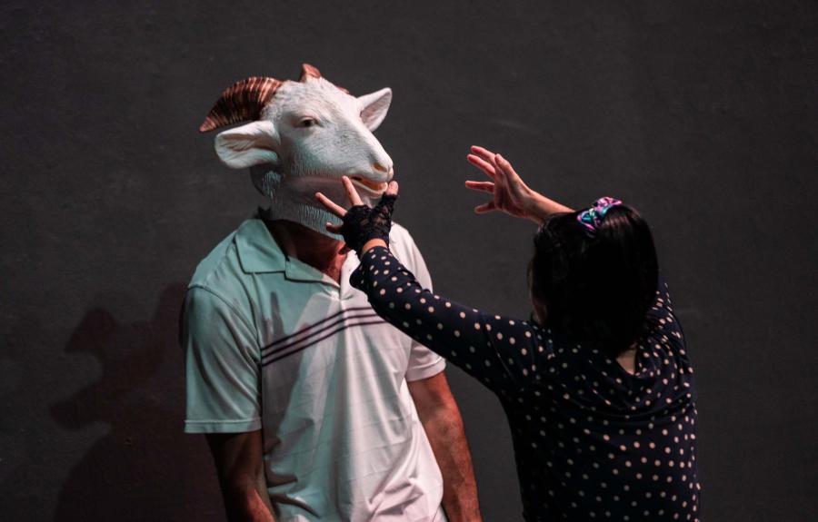 On left a person wearing a goat mask stands still. On right a person reaches towards the face of the person in the goat mask.