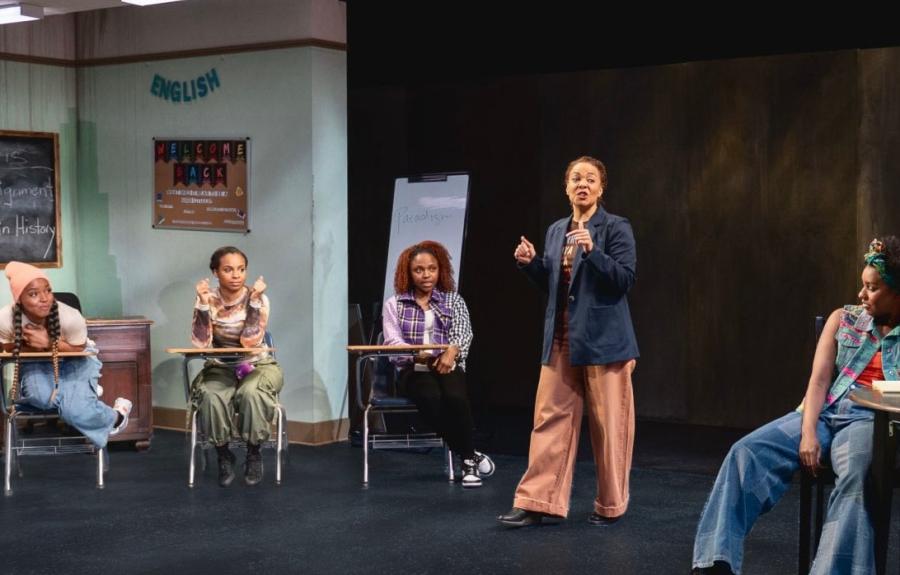 Six actors are on stage during a production of FISH, including a teacher who is standing up and speaking, and five students sitting at classroom desks. In the background there is banner on the wall that says English, a map of the United States, a poster that says Welcome Back, a standing dry-erase board with the word Paradigm written on it, and a chalkboard with writing that says Ms. Harris English Assignment: Black Woman in History.