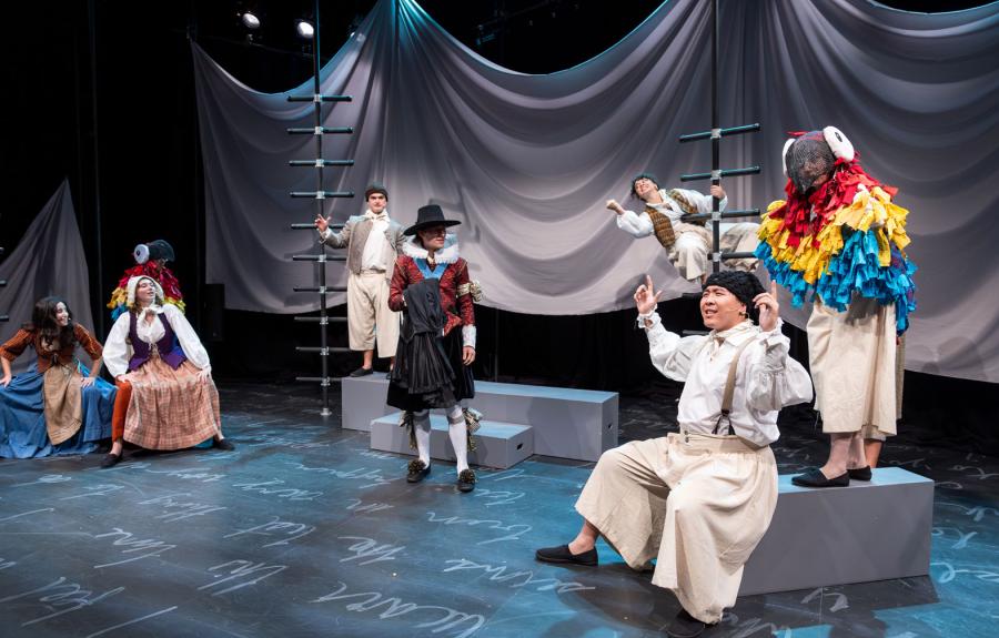 An ensemble of performers scattered across the stage, sitting and standing, on a stage set with black ladders, gray steps and podiums, a sheet made to look like water, and white script writing on the ground. 