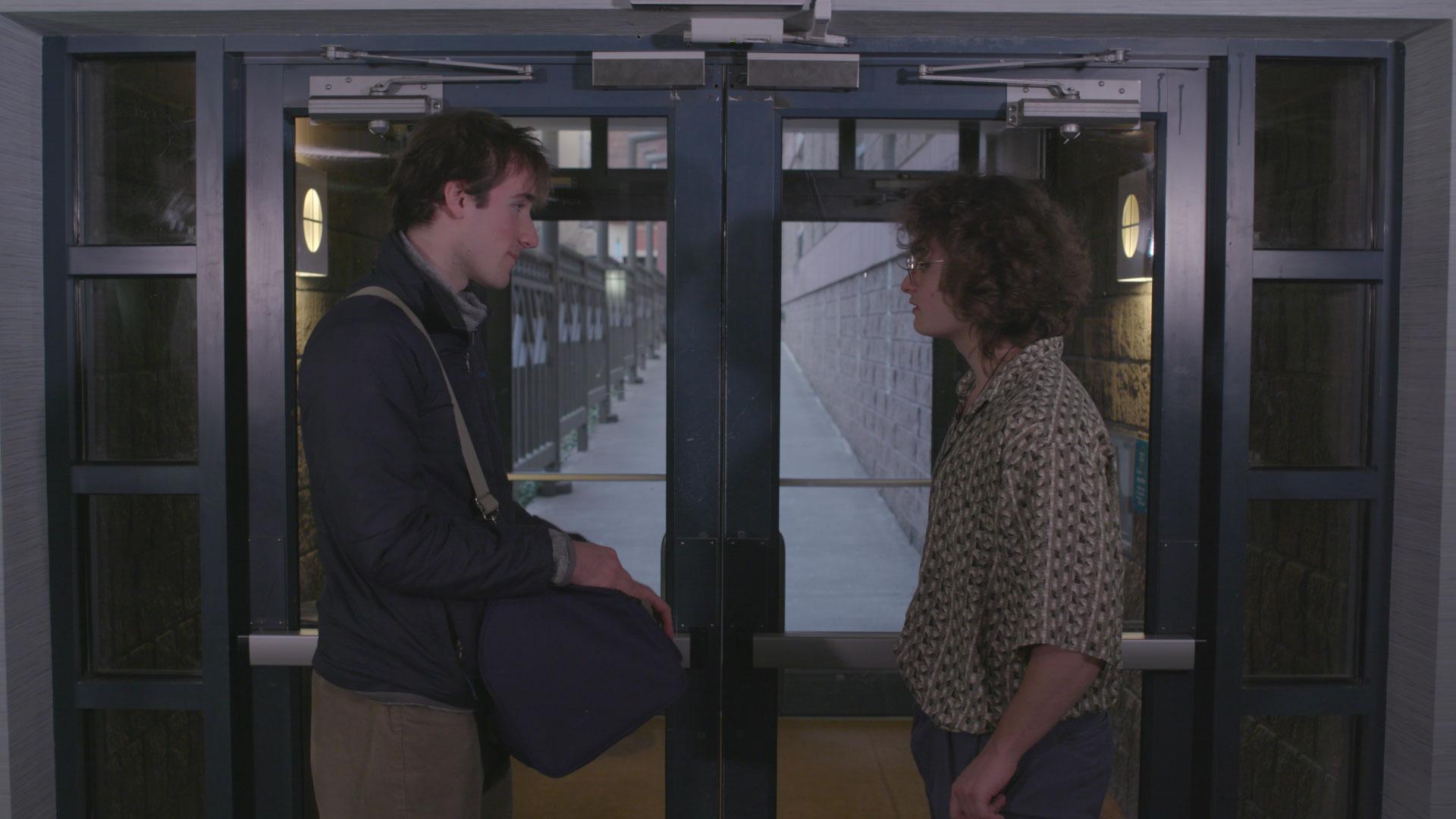 Two young adult men face each other in front of a door.