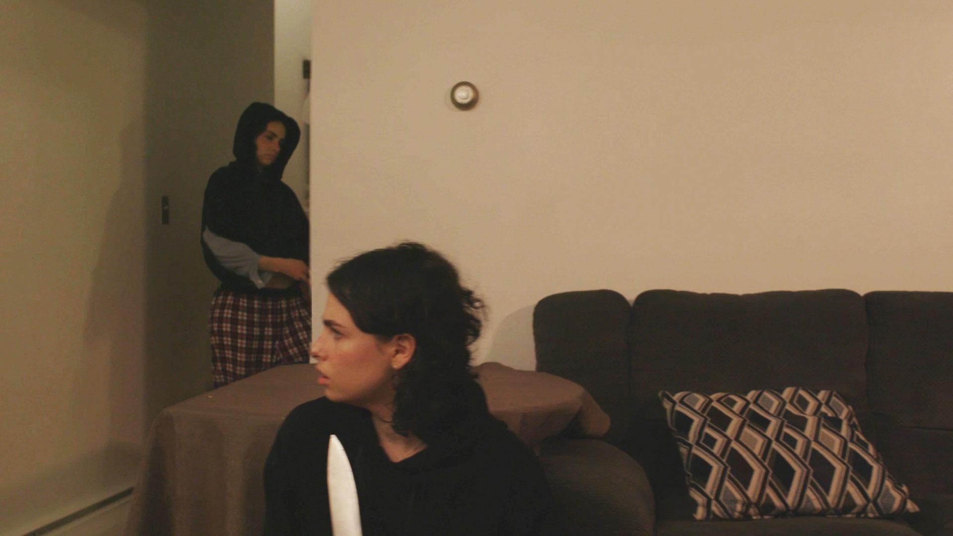 A woman sitting on a couch looks back at another woman opening a door inside an apartment.