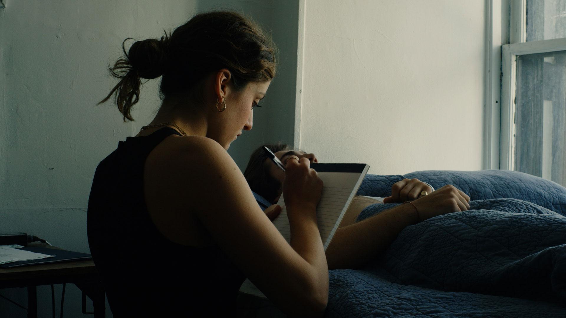 A woman holding a note pad and a pen sitting beside a man laying in bed
