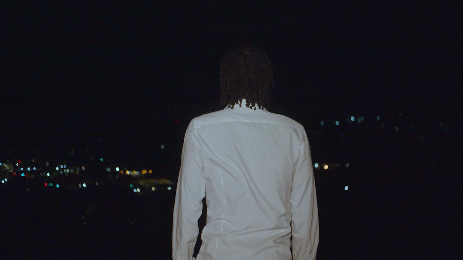 A person in a white shirt staring at the city lights in the distance.
