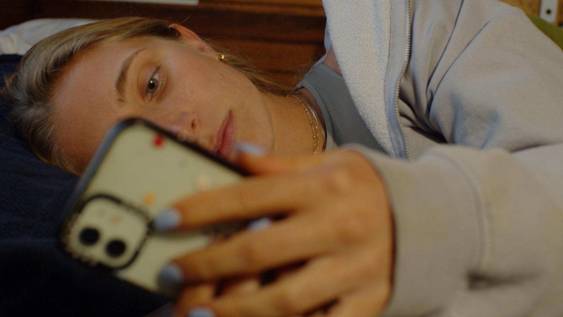 A young woman is holding her phone staring at the screen while lying in bed.