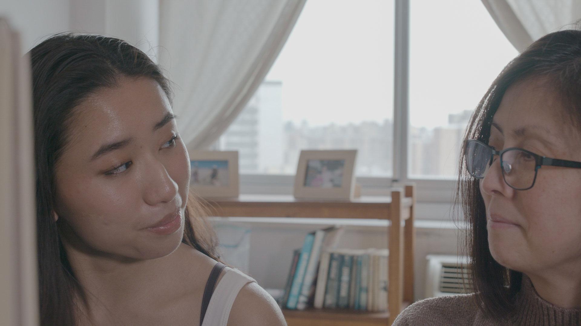 A younger woman and an older woman stand side by side looking at each other inside an apartment.