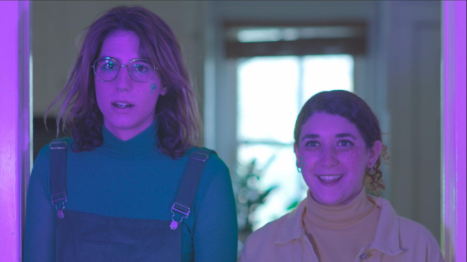 Two people stand next to each other indoors. The person on the right is smiling.