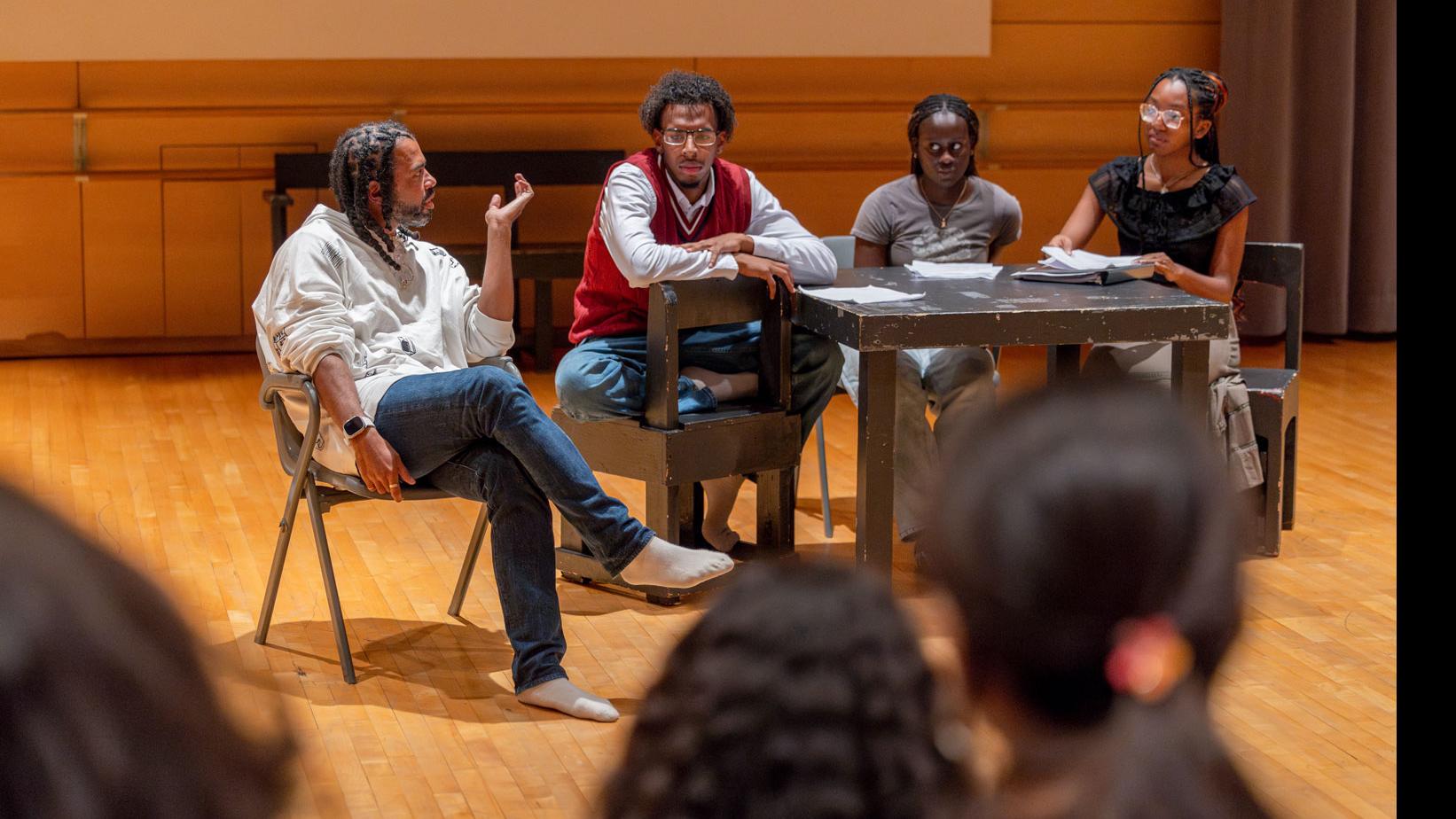 Daveed Diggs sits in a chair on and speaks with three students seated around a table on stage with scripts.