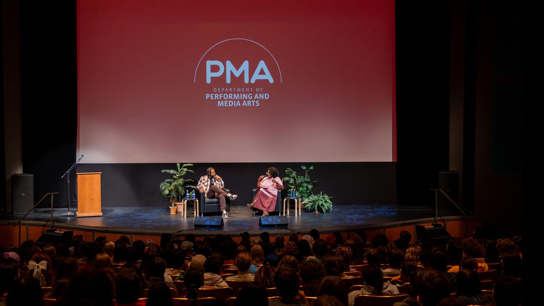 From the middle of the orchestra section of the Kiplinger Theatre. In the foreground, a crowd of people sit in the audience, in the background Daveed Diggs and PMA Department Chair Samantha Noelle Sheppard sit on stage and speak, with a large screen showing the PMA logo.