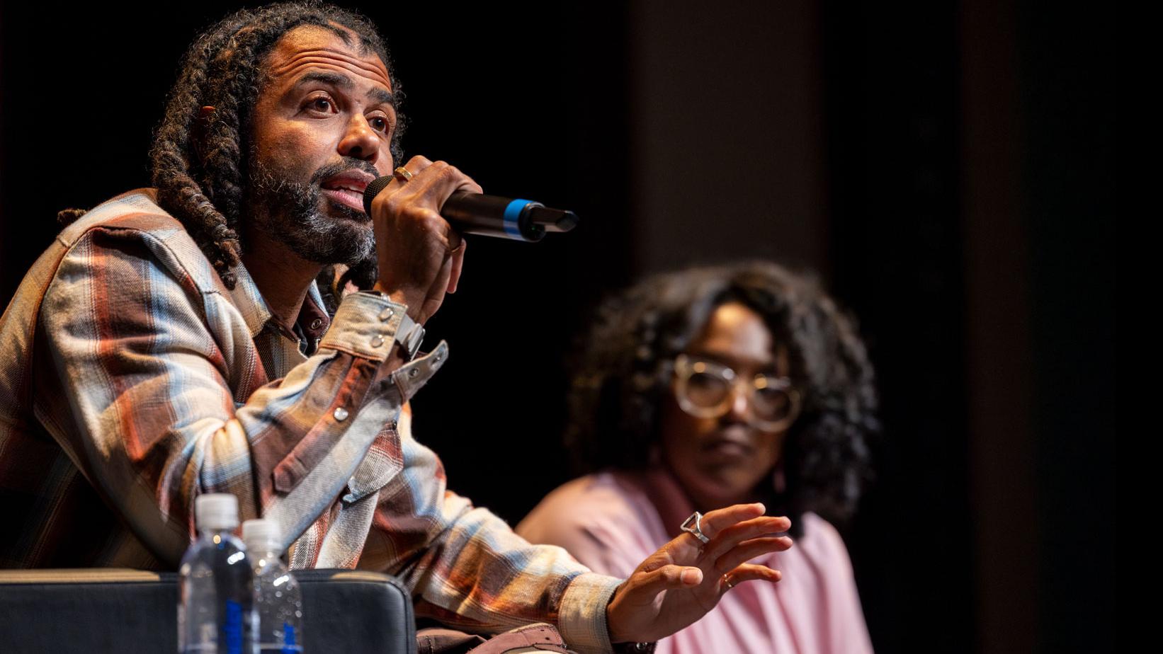 Daveed Diggs sits and speaks into a microphone on stage in the Kiplinger Theatre, while PMA Department Chair Samantha Noelle Sheppard looks on in the background.