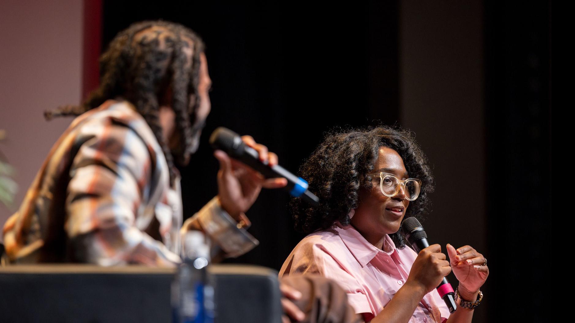 Daveed Diggs sits and speaks into a microphone on stage in the Kiplinger Theatre, while PMA Department Chair Samantha Noelle Sheppard speaks into a microphone in the background.