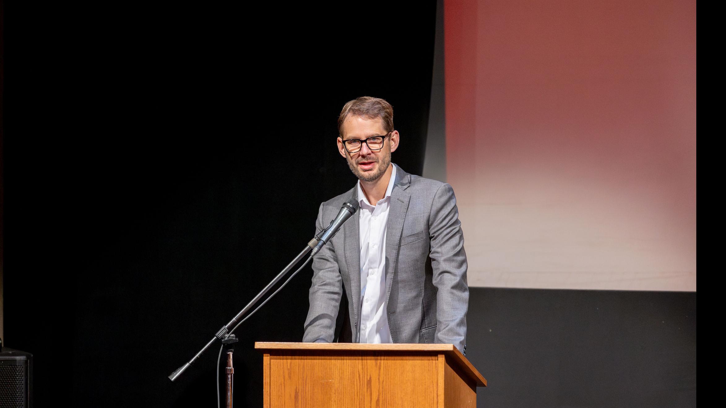 PMA Senior Lecturer Theo Black stands at a podium and speaks into a microphone on stage in the Kiplinger Theatre.