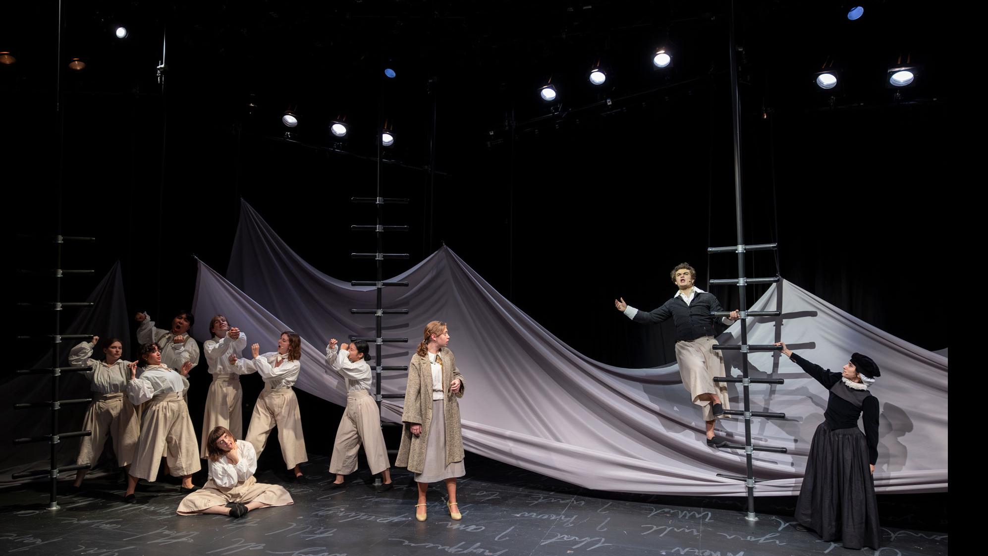 A performer standing on a black ladder, an ensemble of characters with fists up on the left, on a stage set with a sheet made to look like water, and white script writing on the ground.