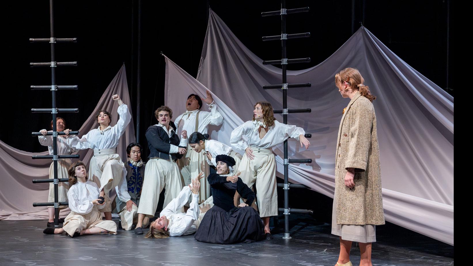 An ensemble of performers on stage, sitting, standing, and leaning on black ladders, with a sheet made to look like water and white script writing on the stage.