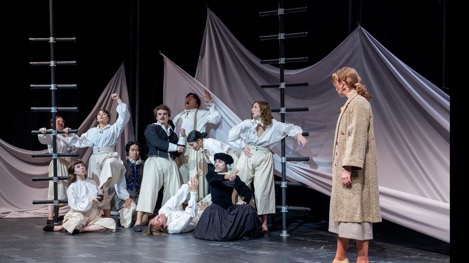 An ensemble of performers on stage, sitting, standing, and leaning on black ladders, with a sheet made to look like water and white script writing on the stage.