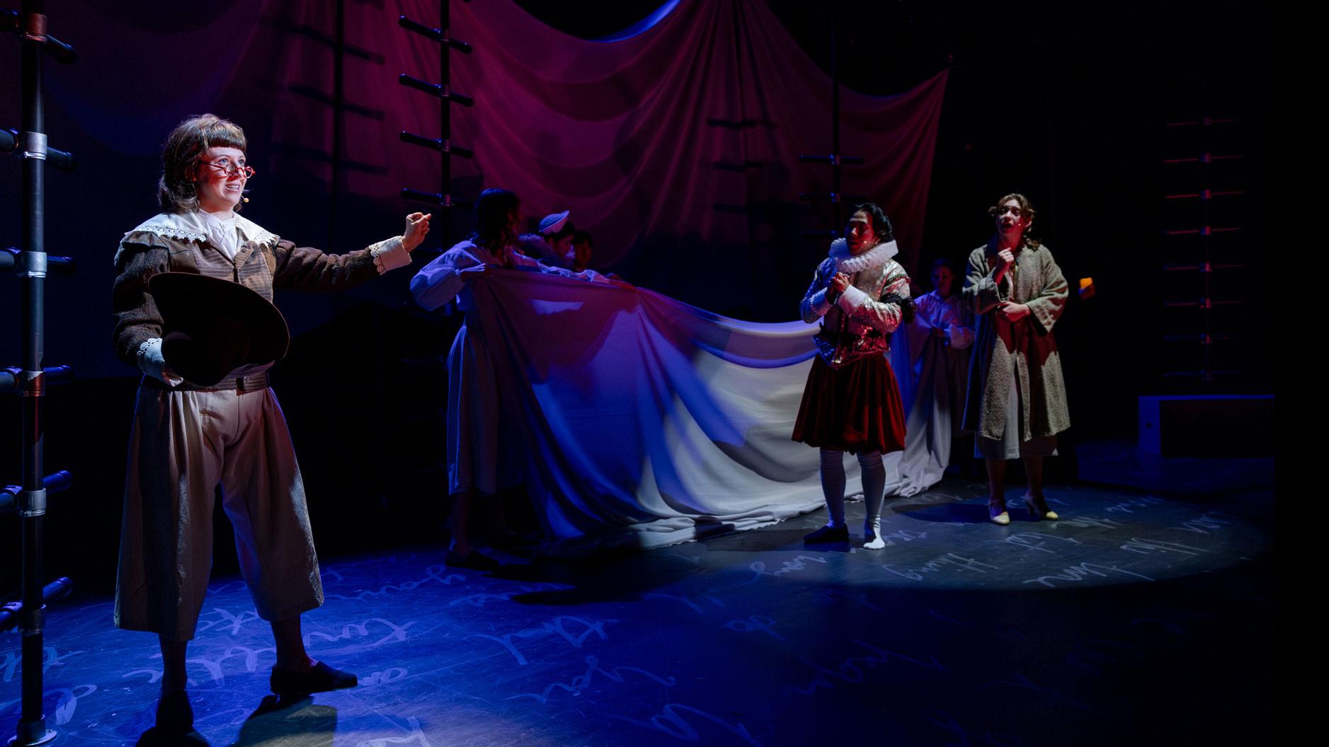 A performer standing with an arm out on a dark stage, while several performers look on in the background. 