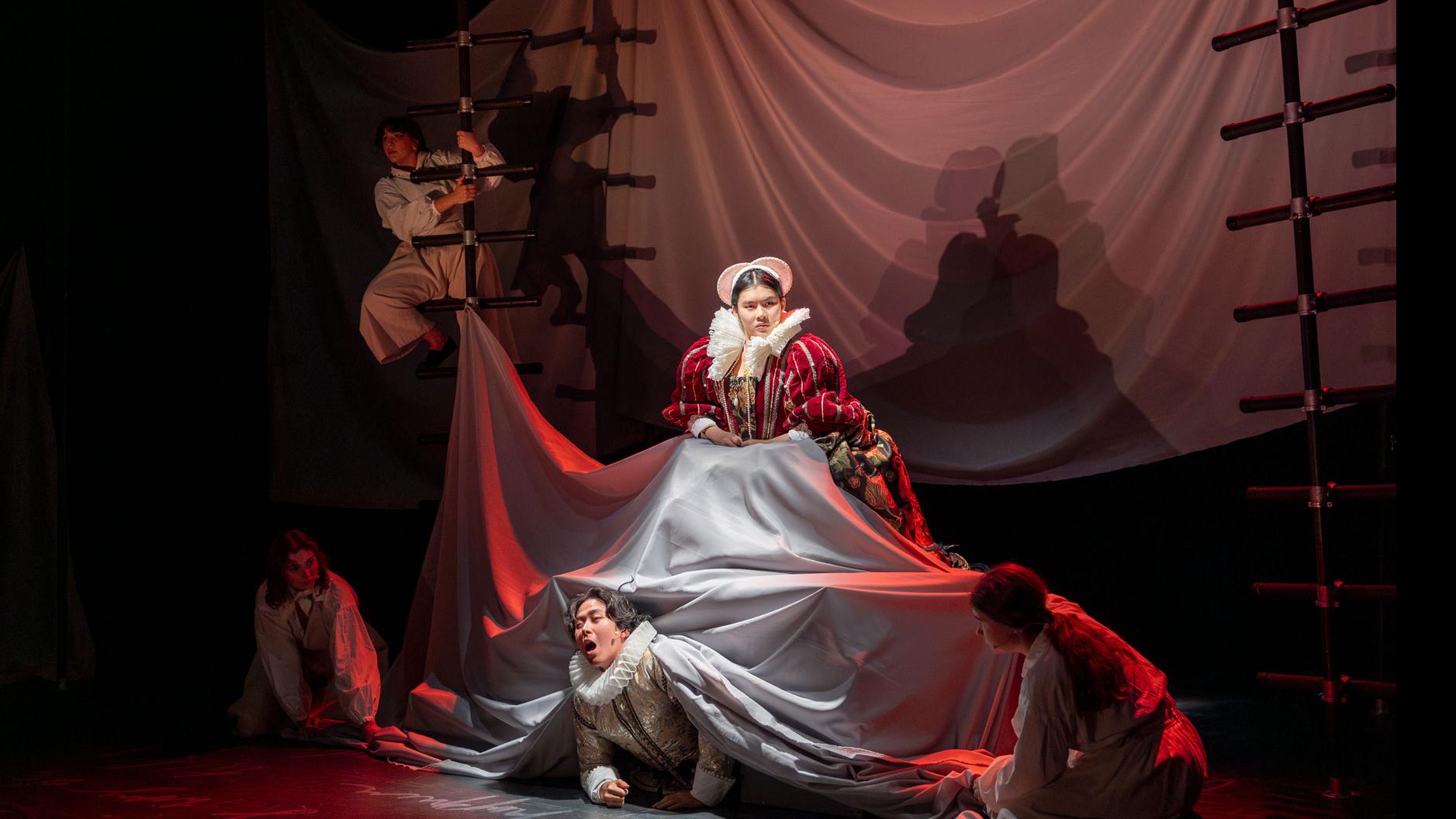 A performer on stage sitting atop a sheet, while another performer emerges from under the sheet, and members of the ensemble on either side, under red light with black ladders.