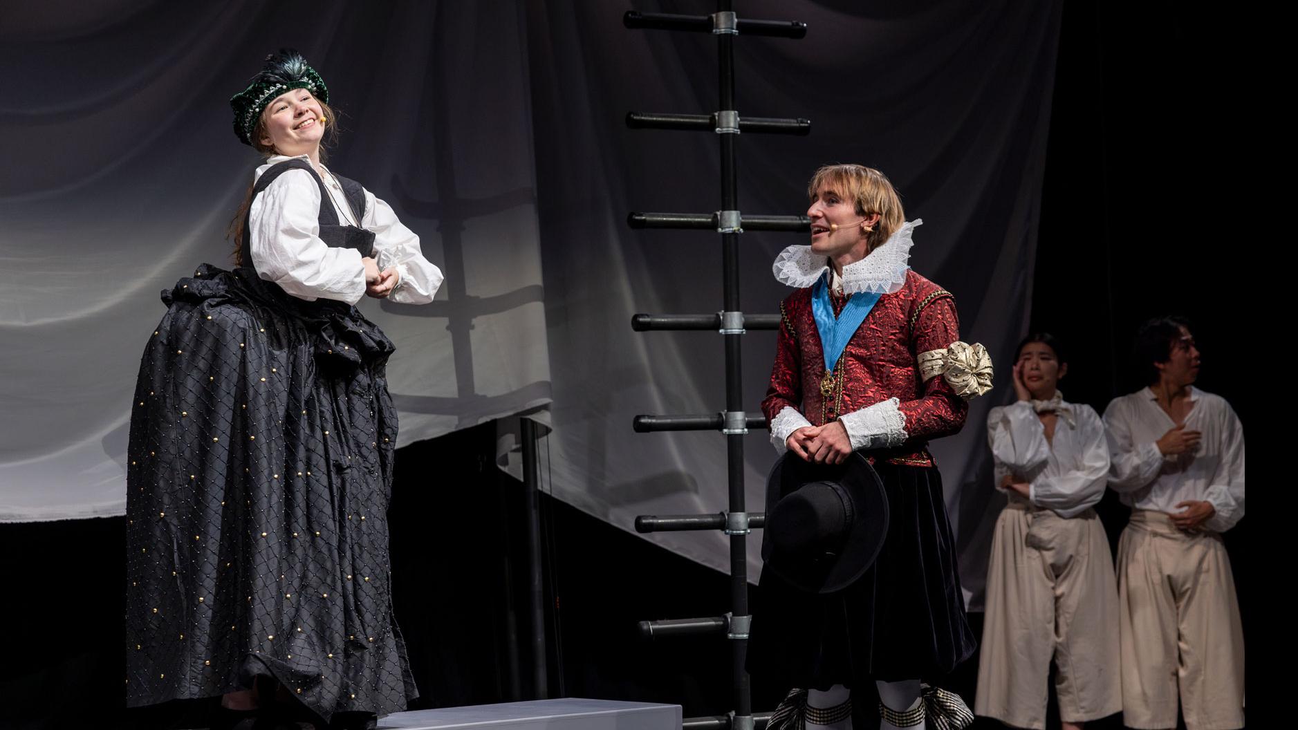 A performer standing on a platform posing, while another performer looks up toward them, with two performers behind a black ladder in the background.