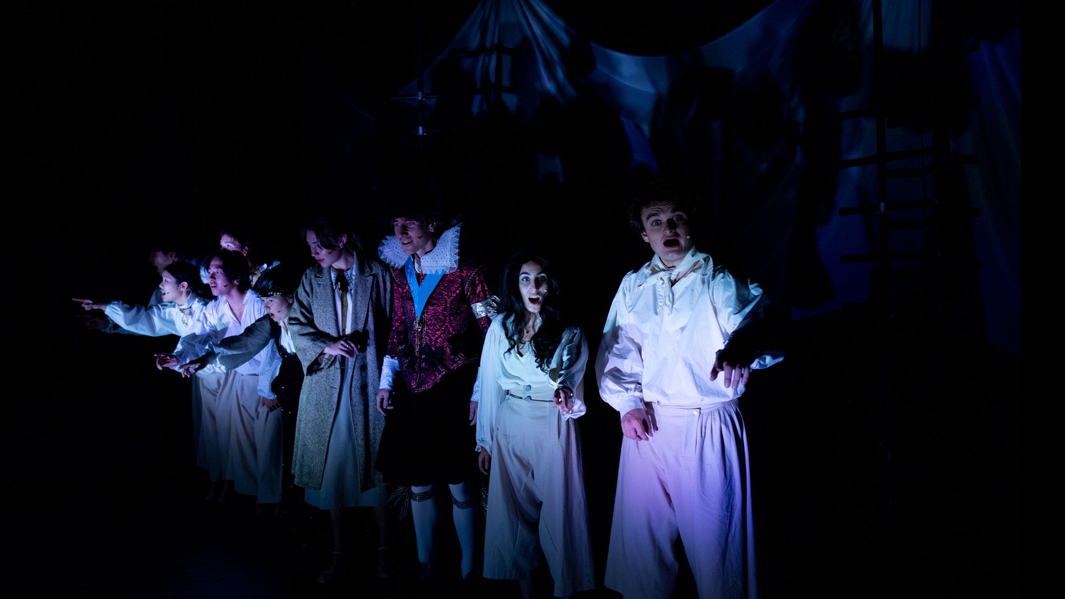 An ensemble of performers on stage in the near dark, standing in a line, looking out and exclaiming. 