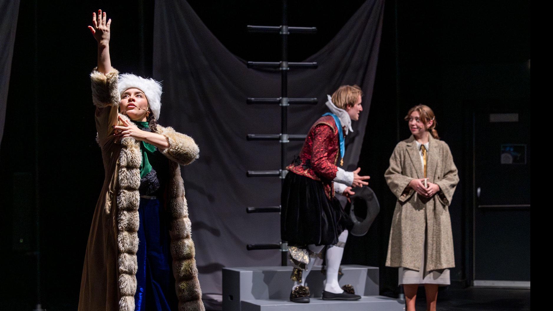 A performer reaching an arm up while two performers behind them speak, on a stage set with black ladders and gray steps.