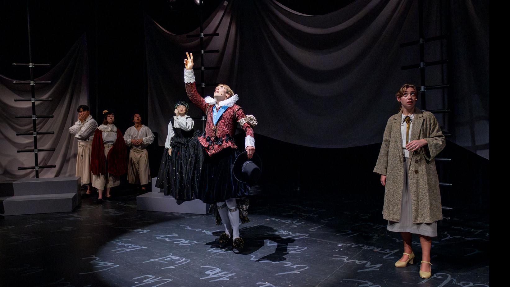 A performer spotlit reaching up toward the ceiling, with an ensemble of performers looking on from the back, on a stage set with black ladders, a sheet made to look like water, and white script writing on the ground.