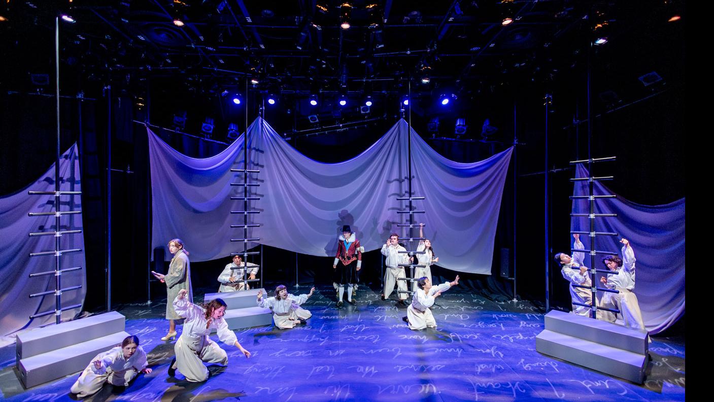 A zoomed out shot of an ensemble of performers scattered across the stage, sitting and standing under blue light, with black ladders, gray steps and podiums, a sheet made to look like water, and white script writing on the ground. 