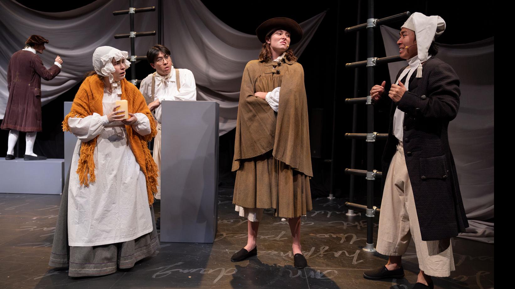 Several performers in a semi-circle conversation, on a stage set with black ladders, gray podiums, a sheet made to look like water, and white script writing on the ground.