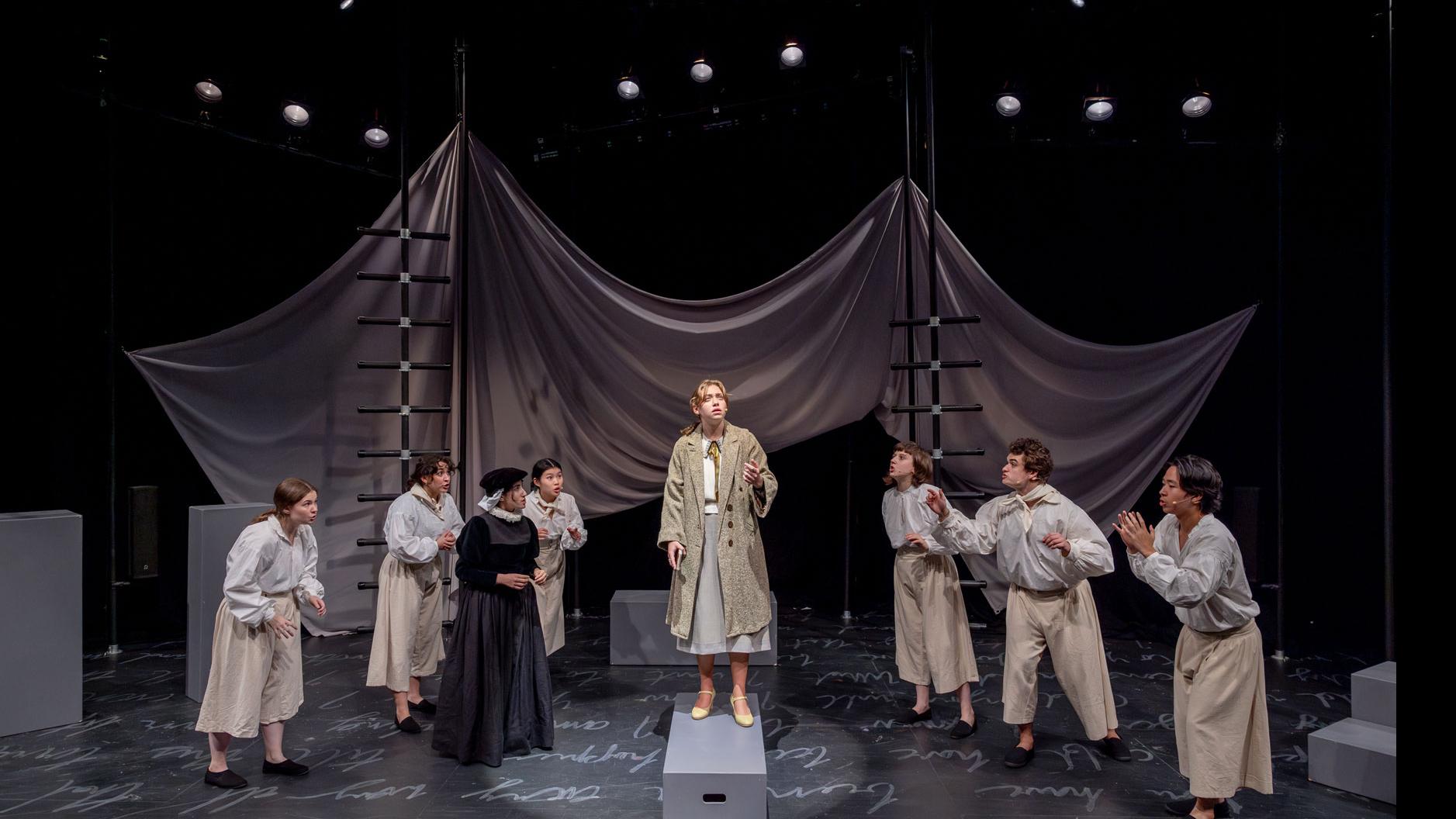 A performer centerstage and spotlit standing on a gray podium looking out, with an ensemble of performers encircling and looking in, on a stage with black ladders, a sheet made to look like water, and white script writing on the ground. 