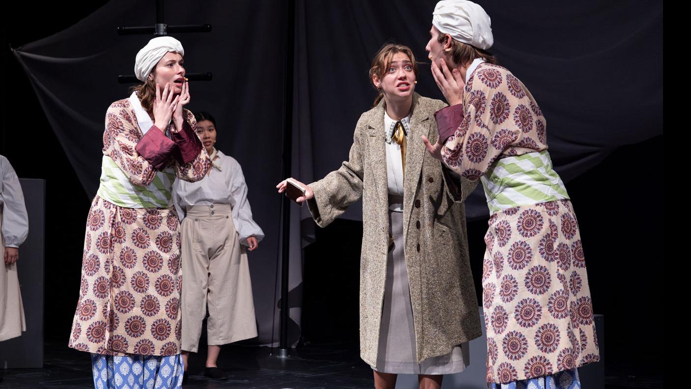 Two performers on stage in identical costumes looking across at each other in mirrored poses, with a performer between them speaking. 