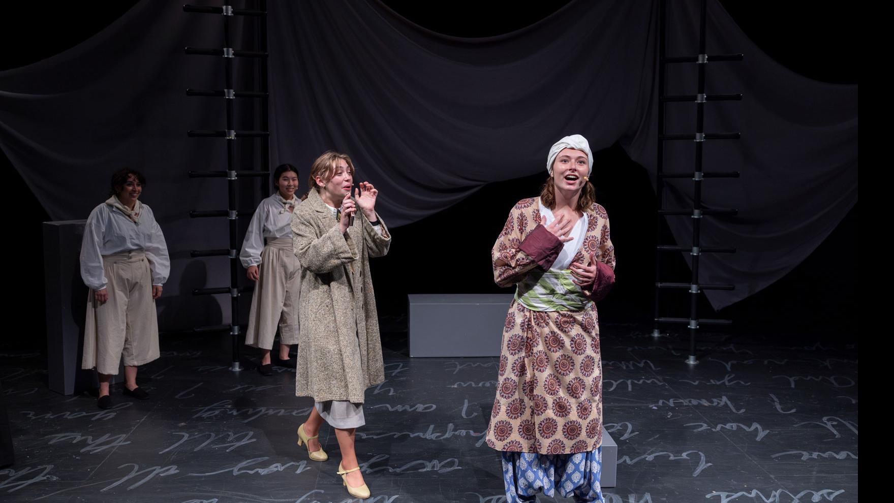 A performer on stage looking out and speaking to the audience, while one performer approaches, and two others stand back left, with white script writing on the ground.