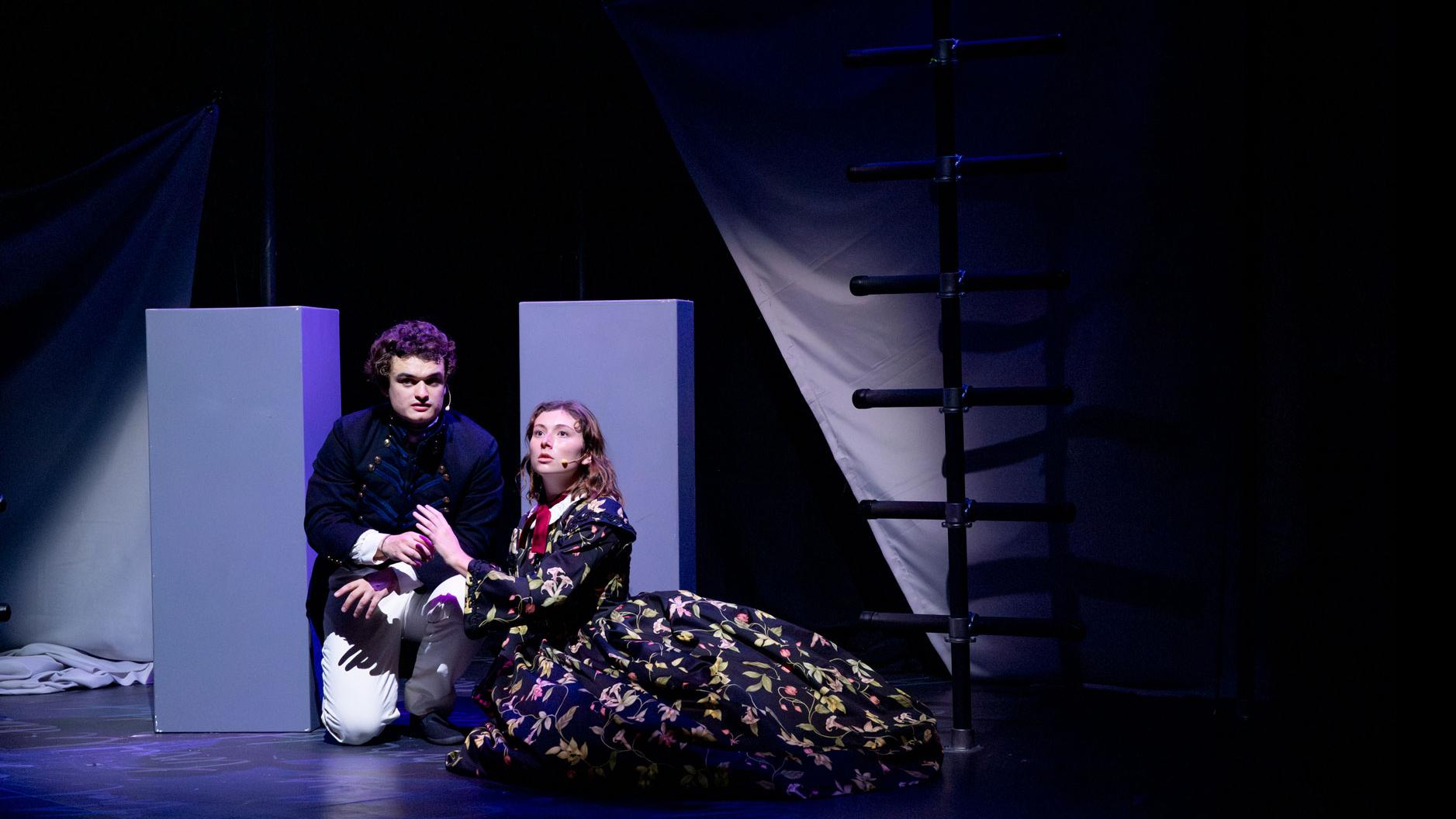 Two performers crouched on the ground with their hands together, looking outward, on a stage set with black ladders and gray podiums.