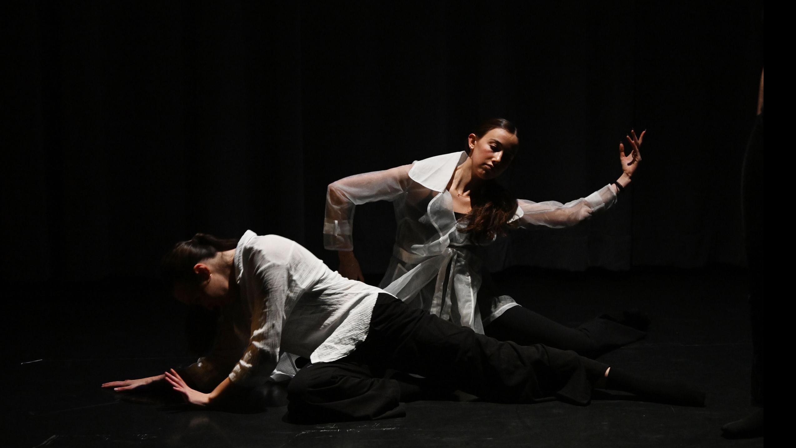 A performer on stage sits on the ground with their head facing down, while another performer sits on the ground and reaches an arm up to the side, in the Class of ’56 Dance Studio Theatre