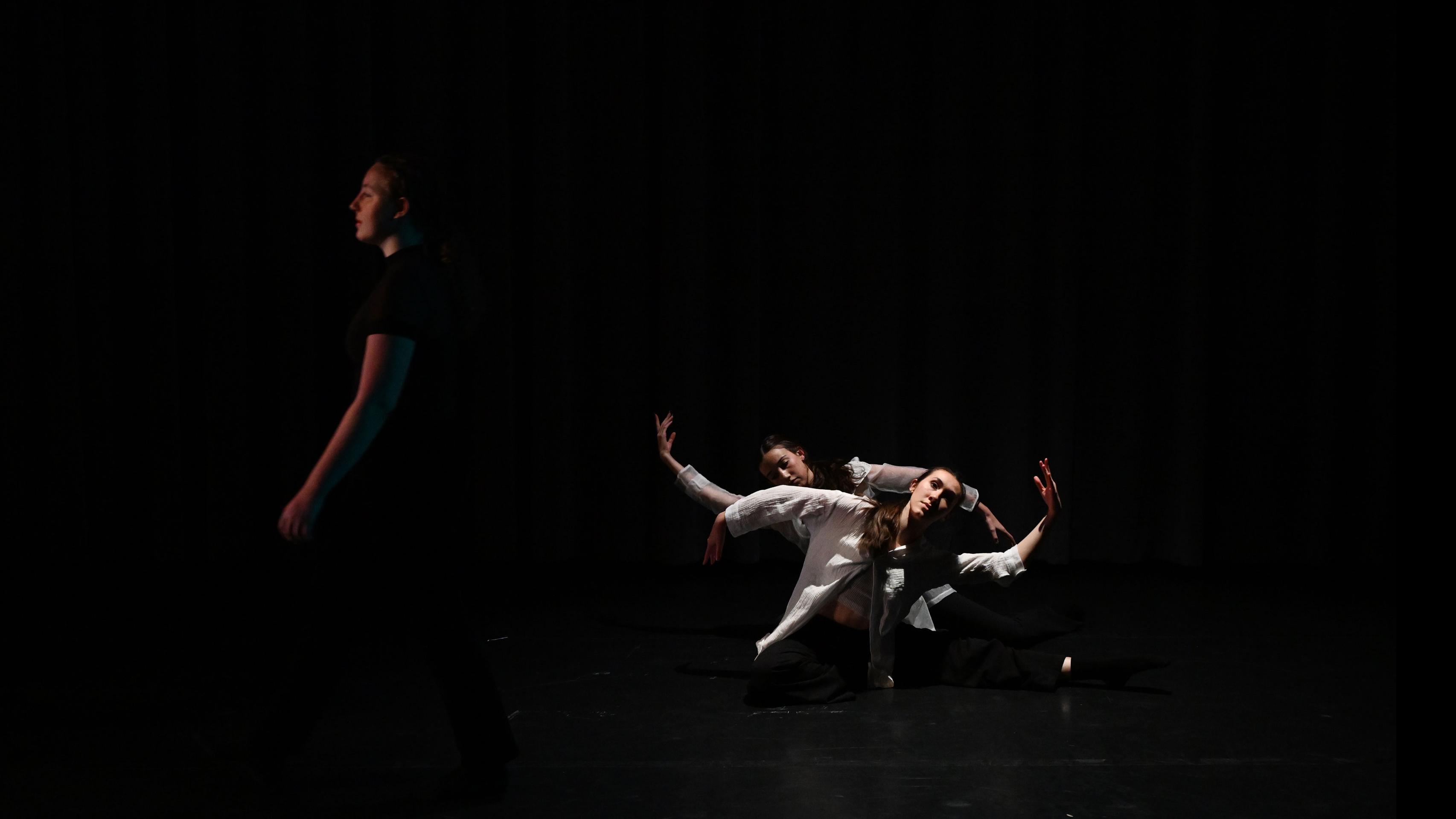 A performer on stage walks to the left, and two other performers sit on the ground, each with an arm raised to the opposite side, in the Class of ’56 Dance Studio Theatre.