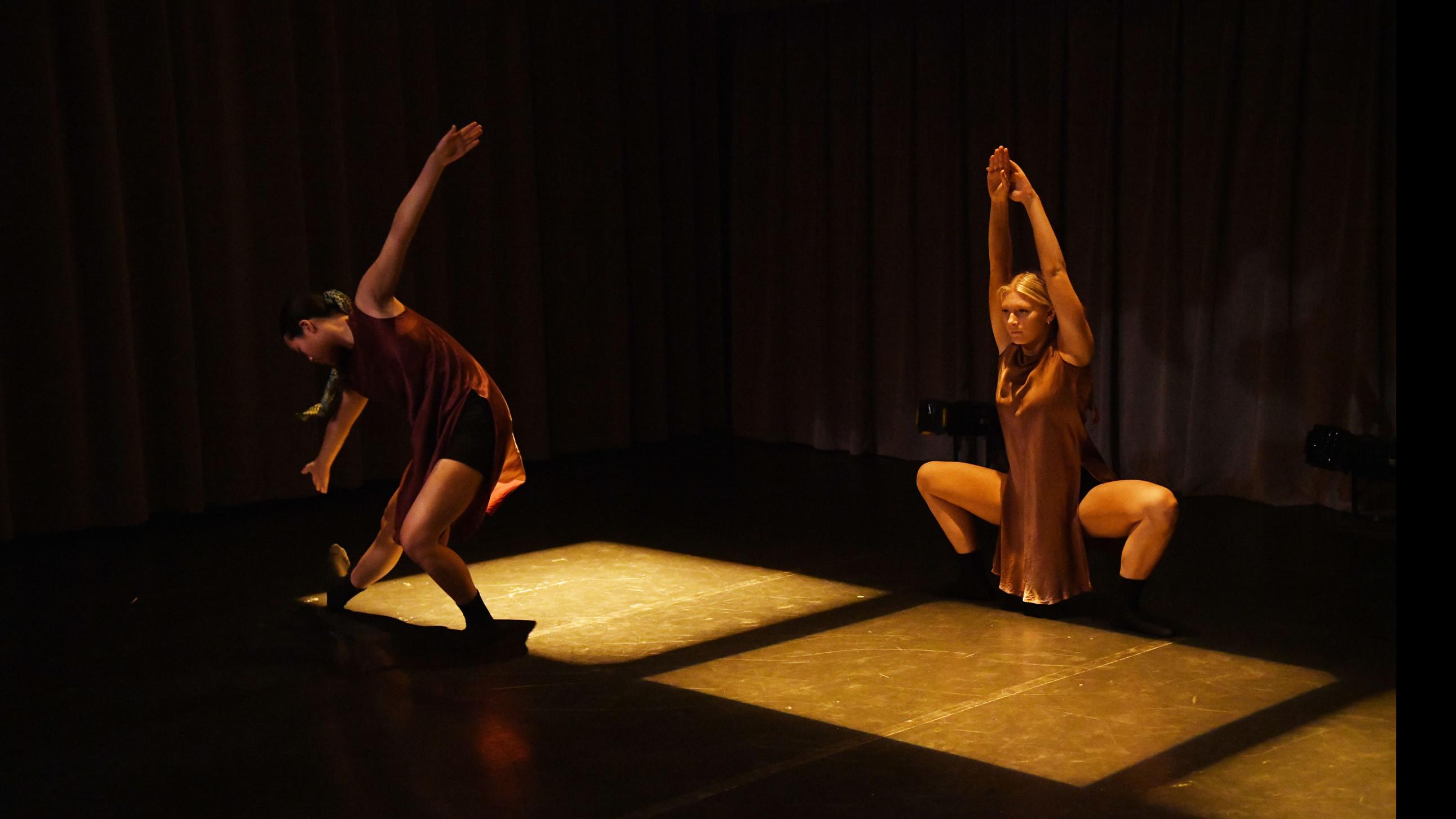 A performer on stage reaches towards their toes, and another performer crouches with hands raised in three squares of light, in the Class of ’56 Dance Studio Theatre.