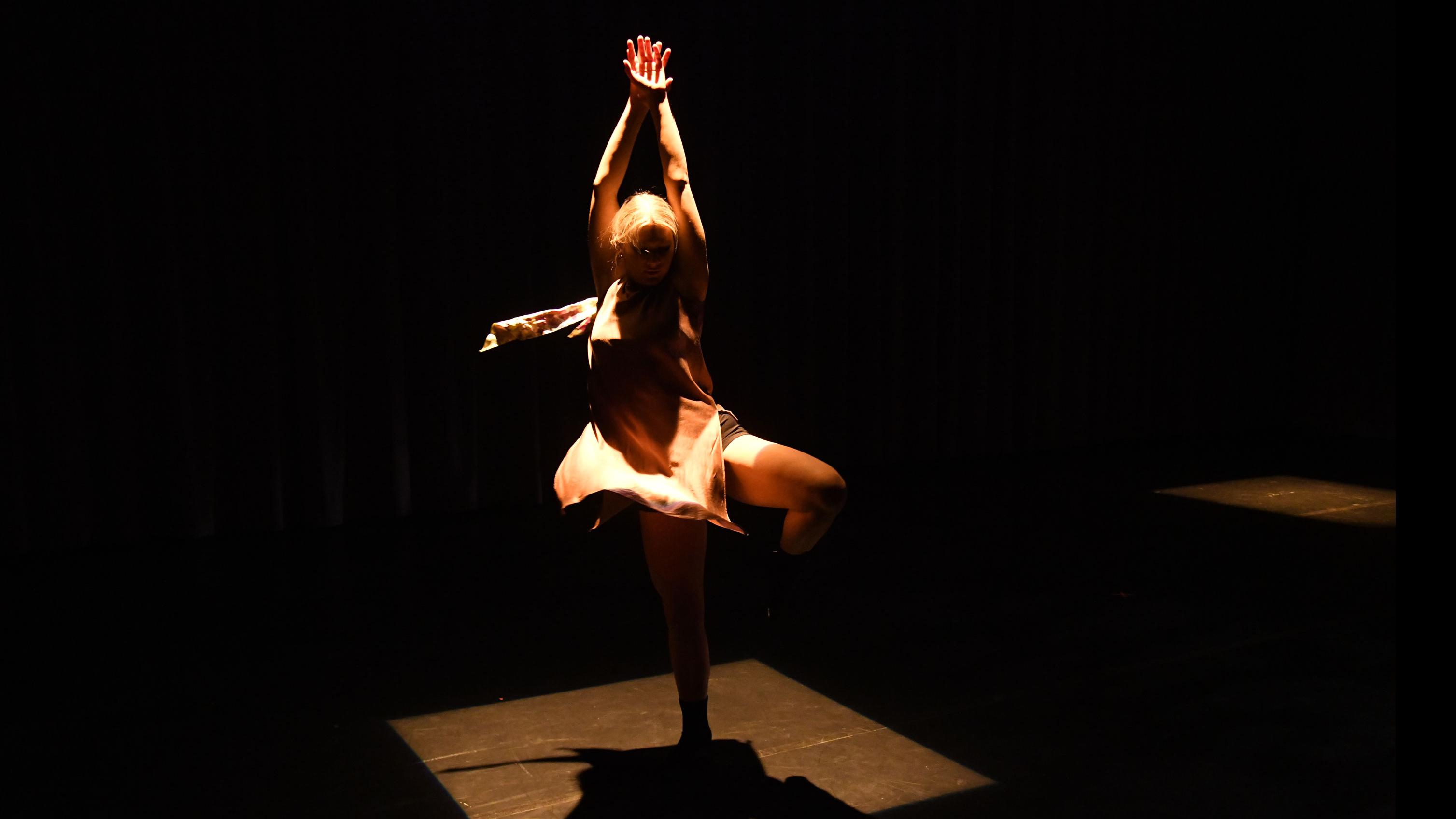 A performer on stage in a square of light in a pirouette pose, in the Class of ’56 Dance Studio Theatre.