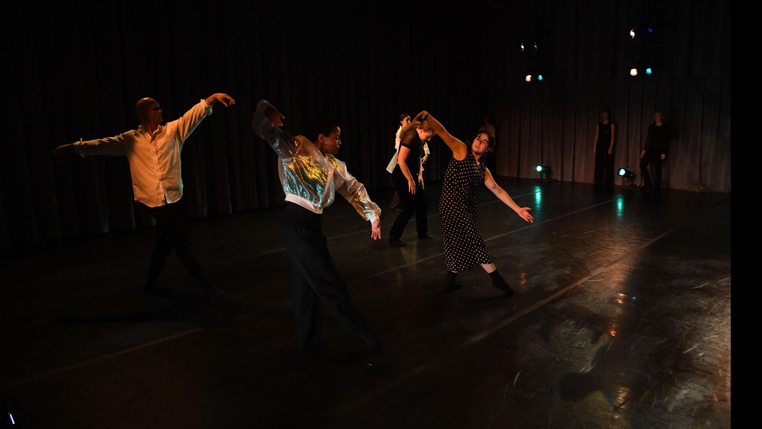 In the foreground, five performers on stage reach their arms out to the side or press their hands to their legs, while two other performers in the background stand against a curtain, in the Class of ’56 Dance Studio Theatre.