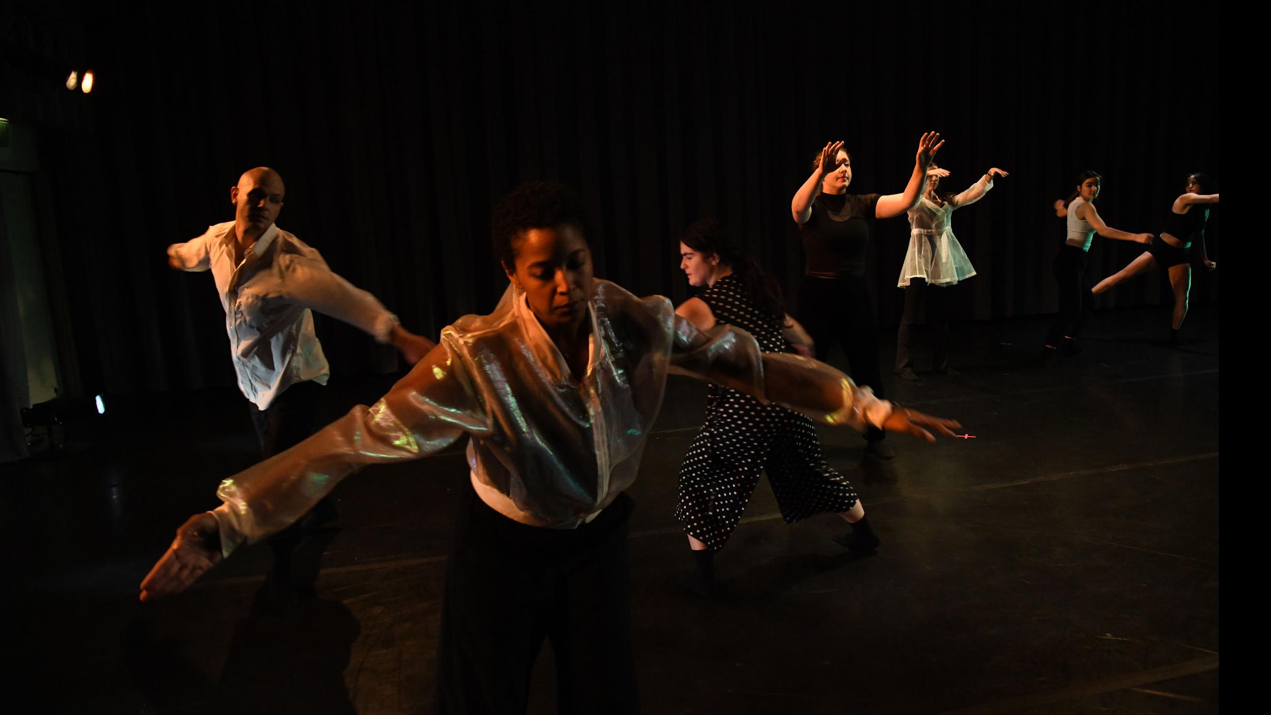 In the foreground, a performer on stage reaches their arms out to the side, with an ensemble of performers in the background in various movement poses, in the Class of ’56 Dance Studio Theatre. 