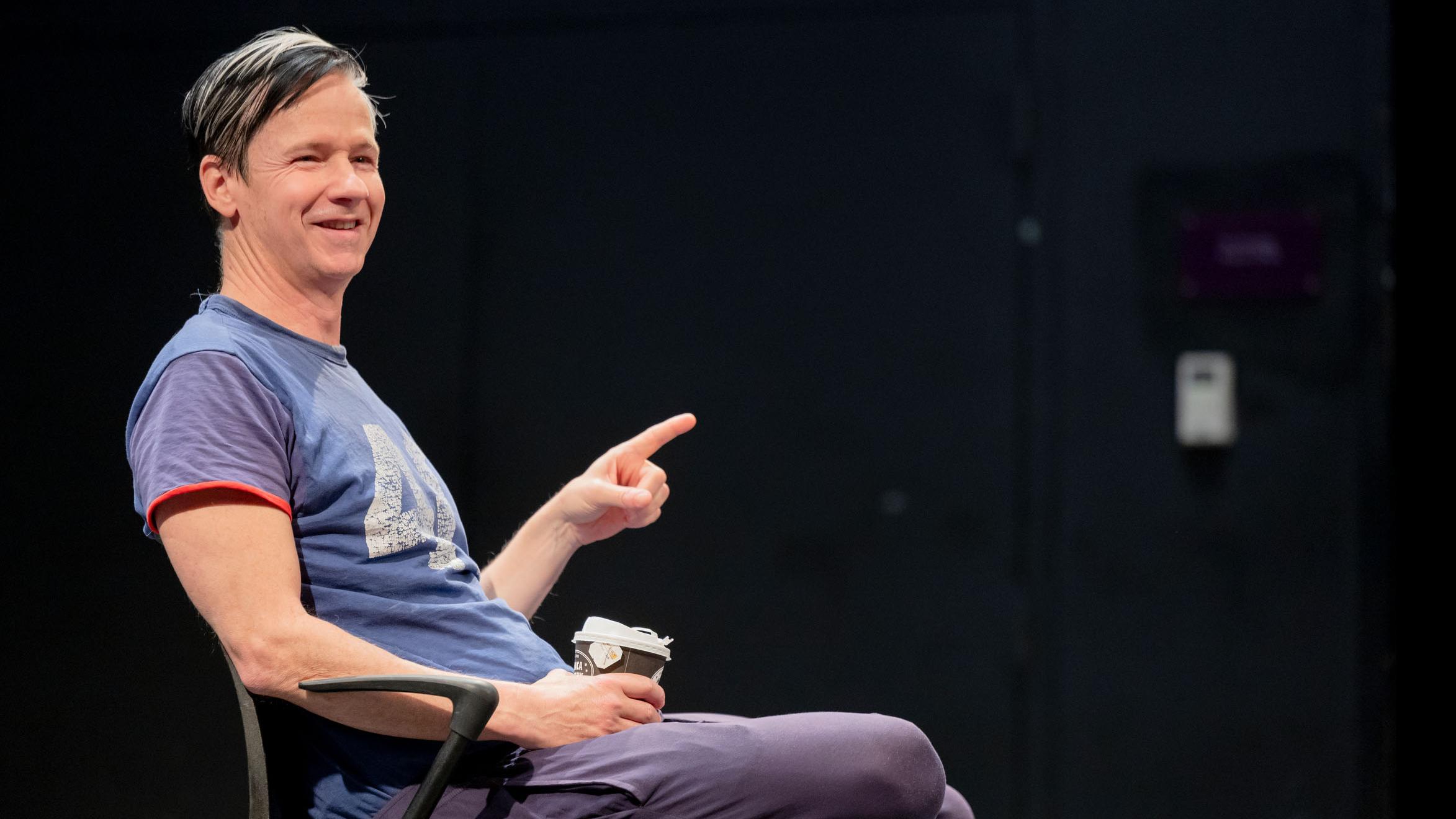 John Cameron Mitchell sits on stage in the Black Box Theatre, smiles and points. Photo credit: Simon Wheeler