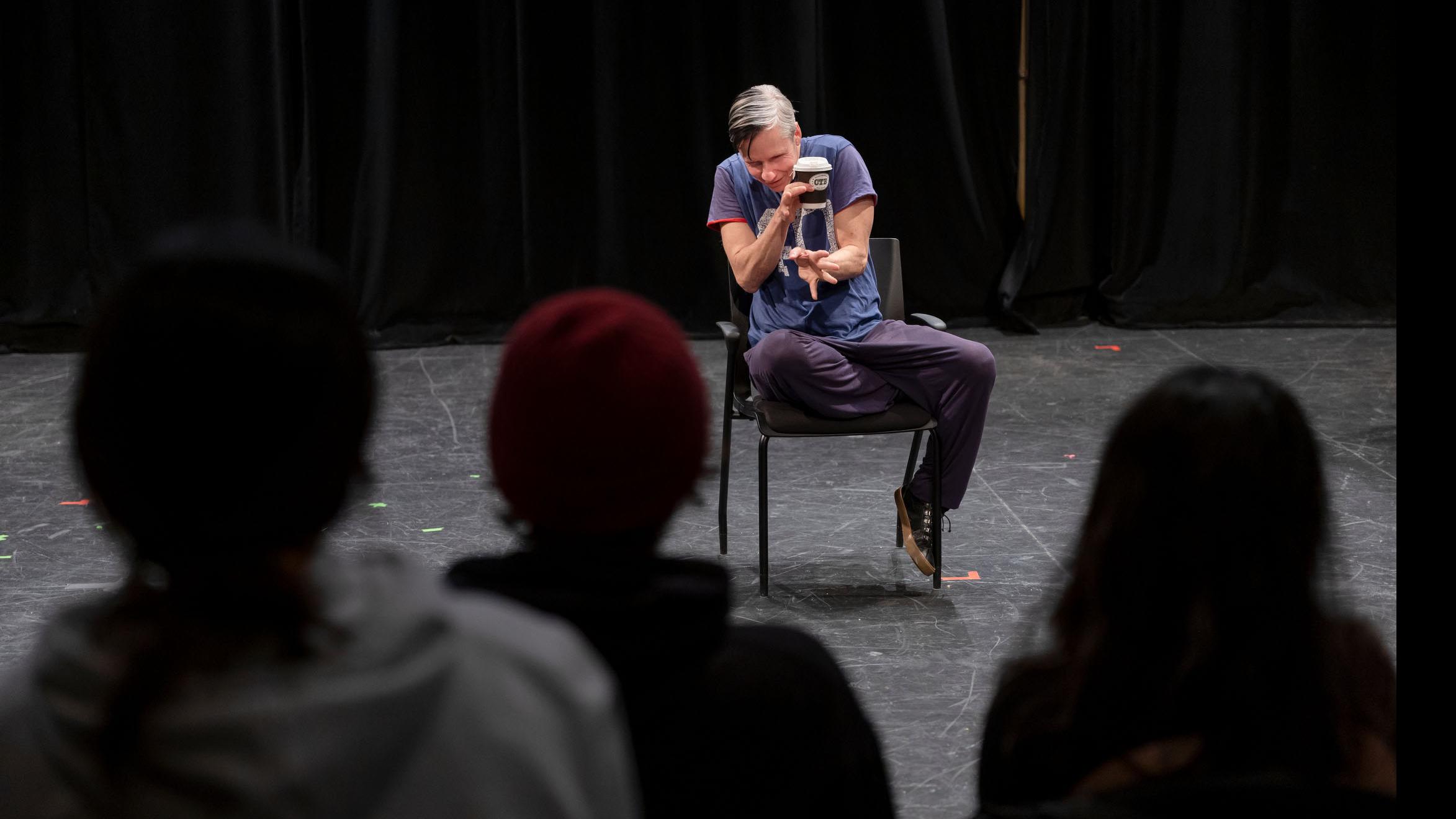 Taken from behind three students in the audience, John Cameron Mitchell sits on stage in the Black Box Theatre and makes a gesture with his hands. Photo credit: Simon Wheeler