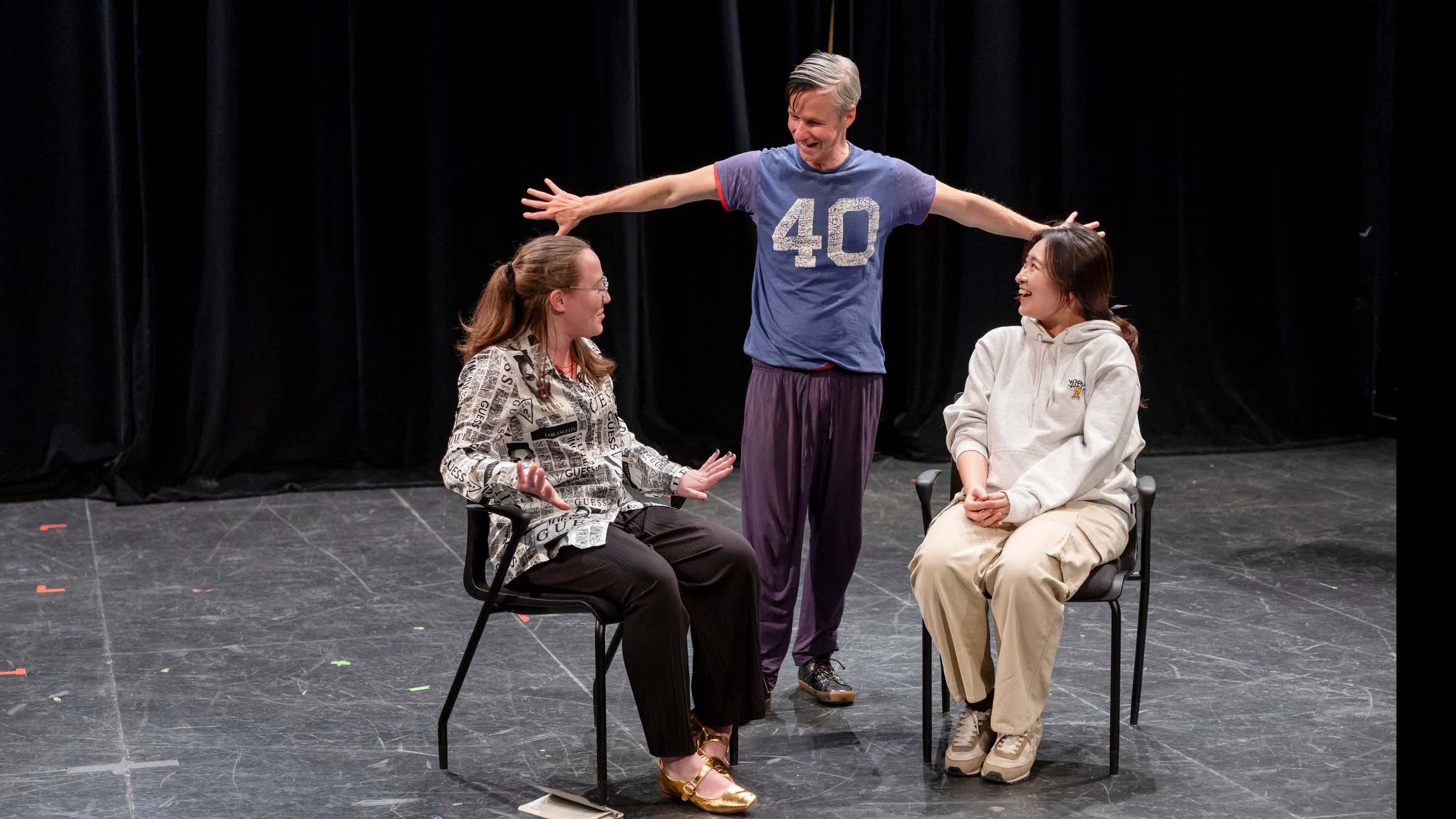 John Cameron Mitchell stands on stage in the Black Box Theatre and speaks to two students seated on stage with notebooks. Photo credit: Simon Wheeler