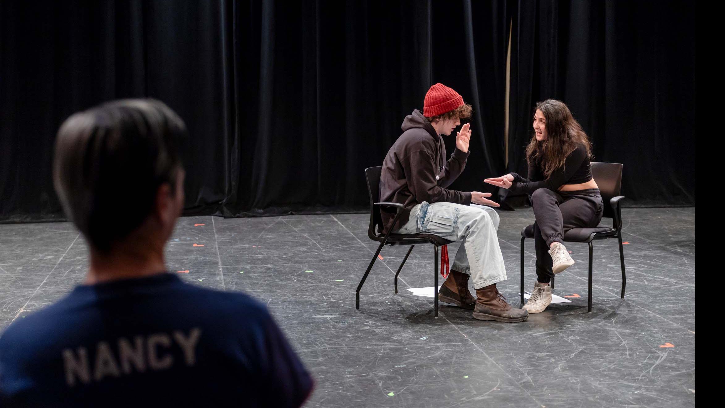 In the foreground, John Cameron Mitchell faces away towards two students seated and engaged in a scene on stage in the Black Box Theatre. Photo credit: Simon Wheeler