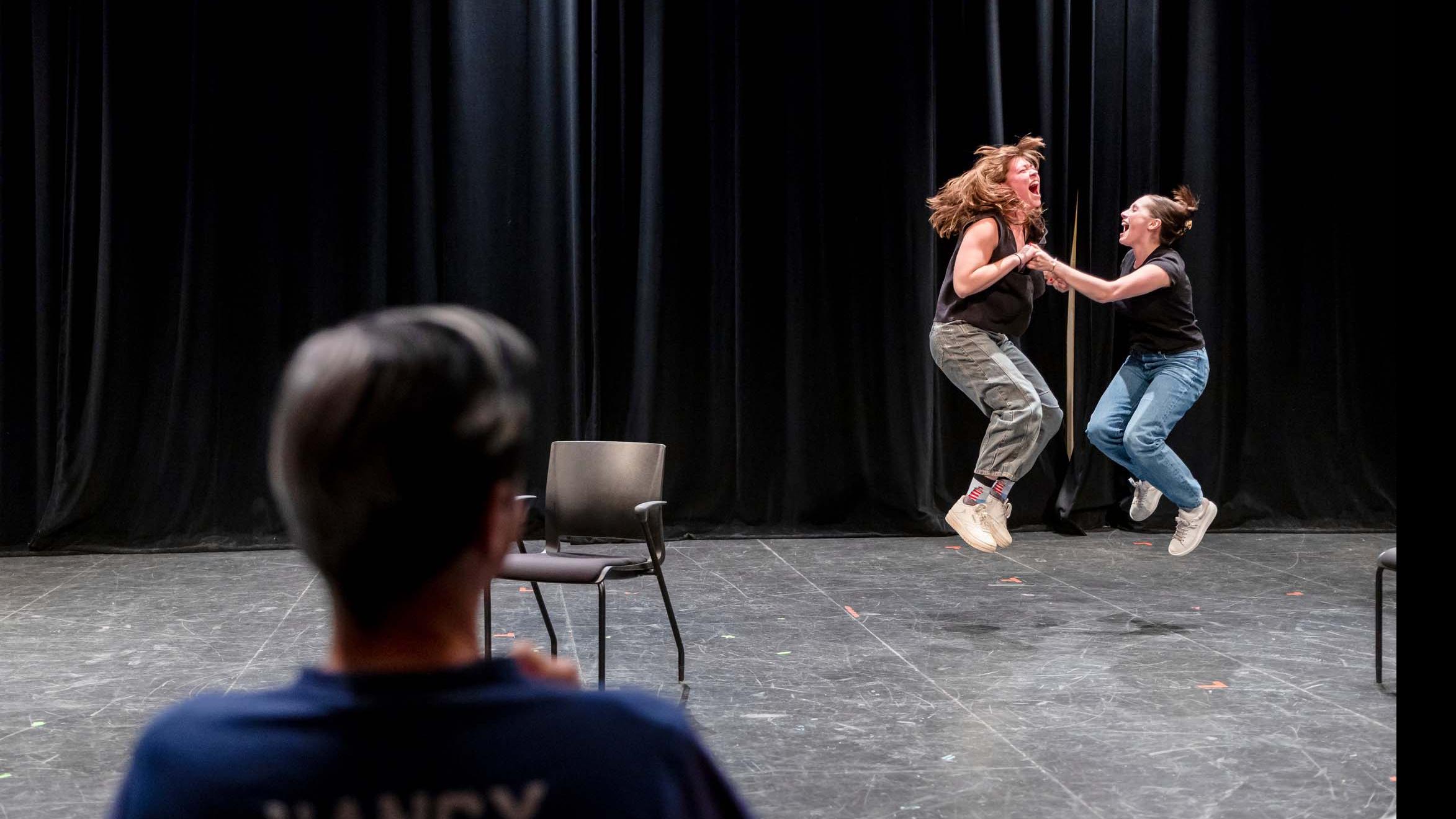 In the foreground, John Cameron Mitchell faces away towards to two students jumping into the air and holding hands in a scene on stage in the Black Box Theatre. Photo credit: Simon Wheeler