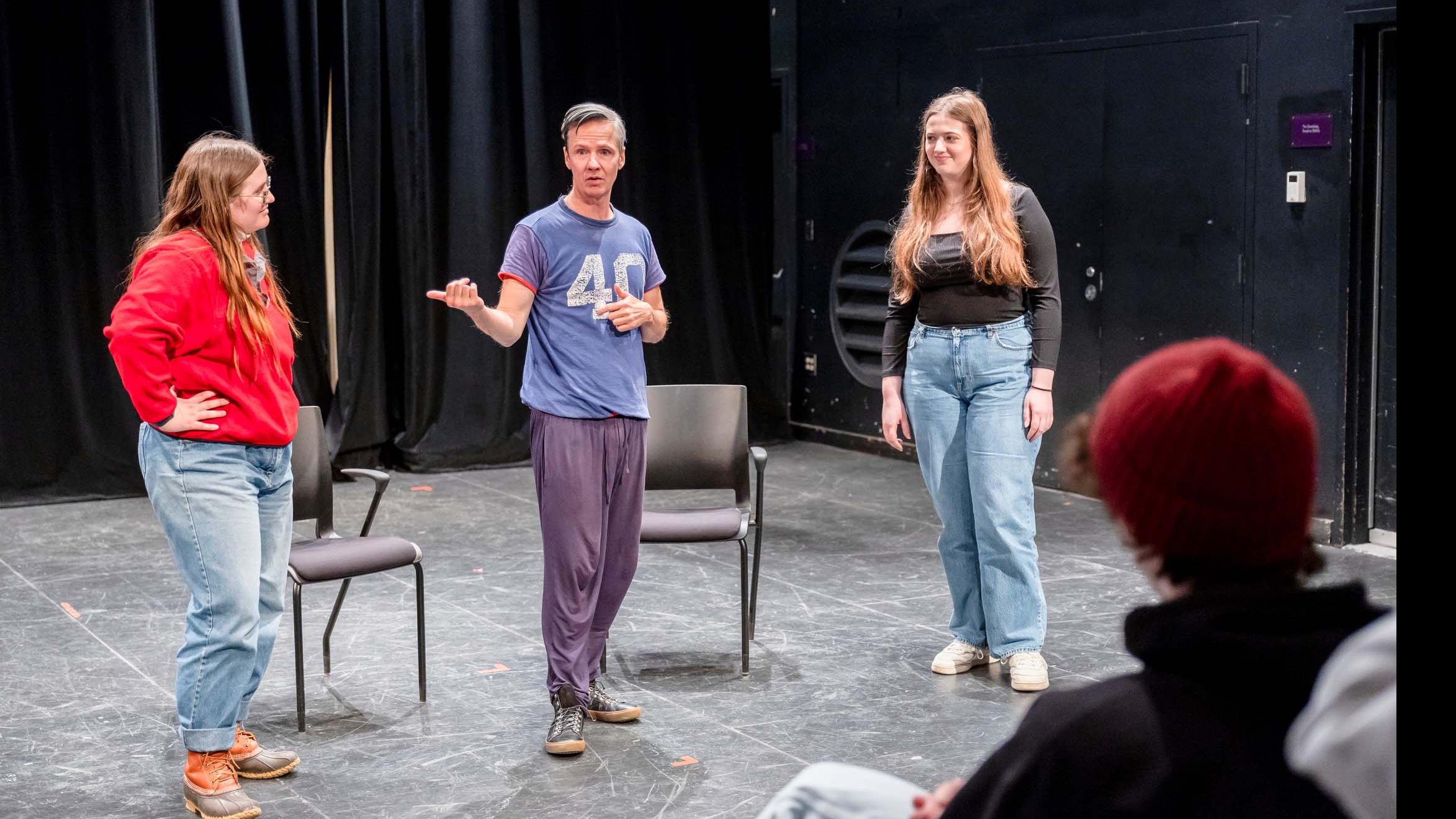 John Cameron Mitchell stands on stage in the Black Box Theatre and speaks to two students standing on stage. Photo credit: Simon Wheeler