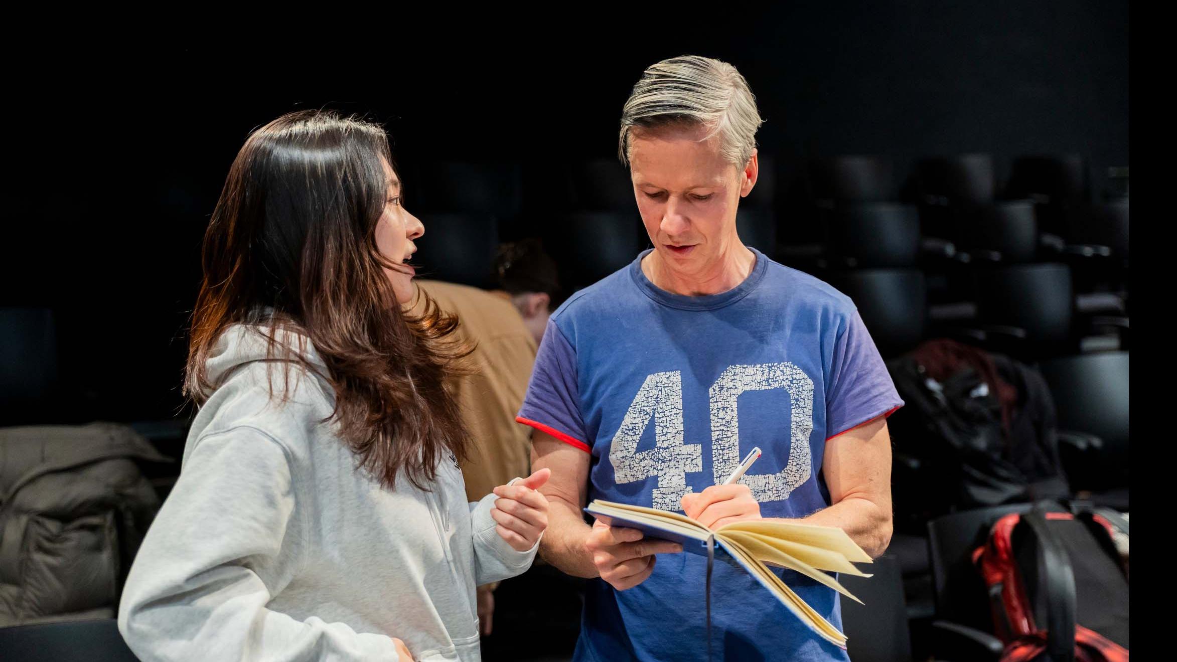 John Cameron Mitchell stands on stage in the Black Box Theatre and writes in a notebook beside a student who speaks to him. Photo credit: Simon Wheeler