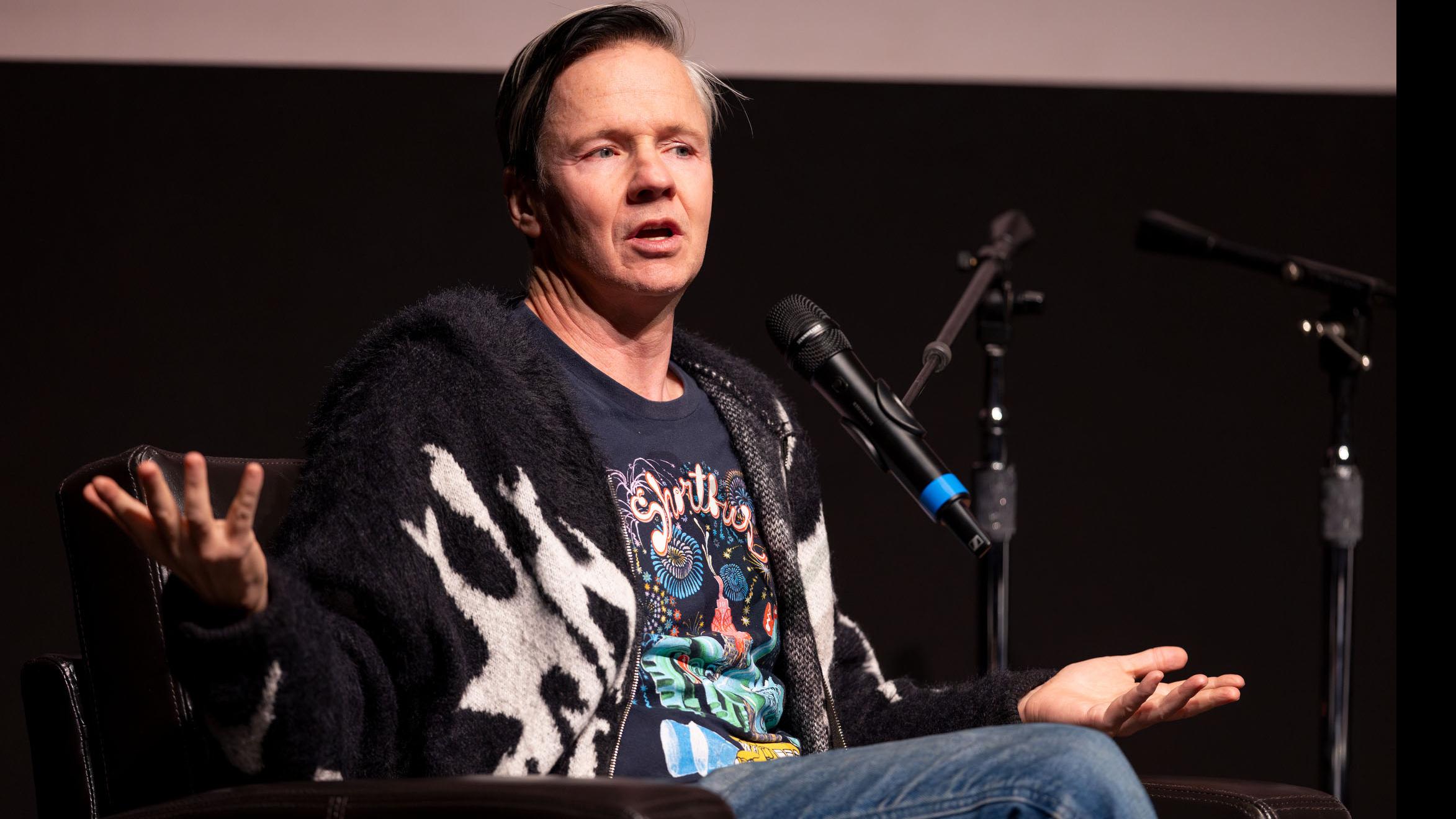 John Cameron Mitchell speaks into a microphone on stage in the Kiplinger Theatre. Photo credit: Simon Wheeler