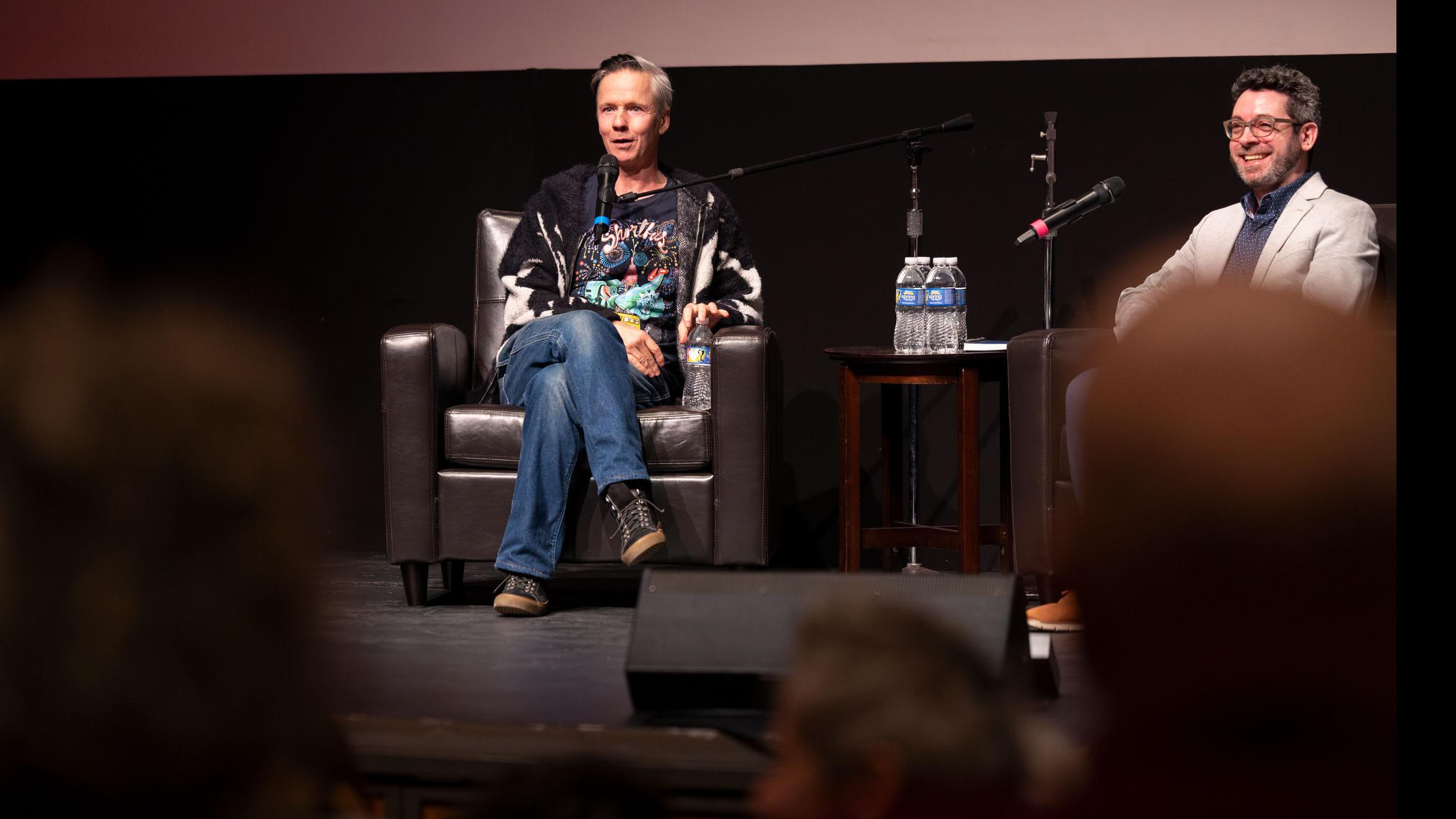 John Cameron Mitchell smiles and looks at the audience while PMA faculty member Nick Salvato smiles, on stage in the Kiplinger Theatre. Photo credit: Simon Wheeler