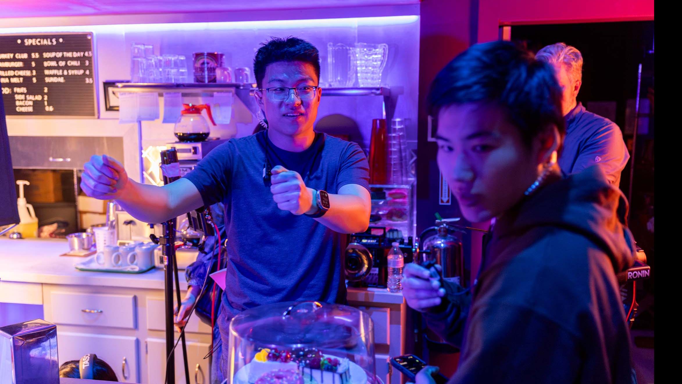Two student crew members talk and gesture from behind the countertop on the set of the PMA Studios short film Milkshake Kiss. Photo credit: Simon Wheeler