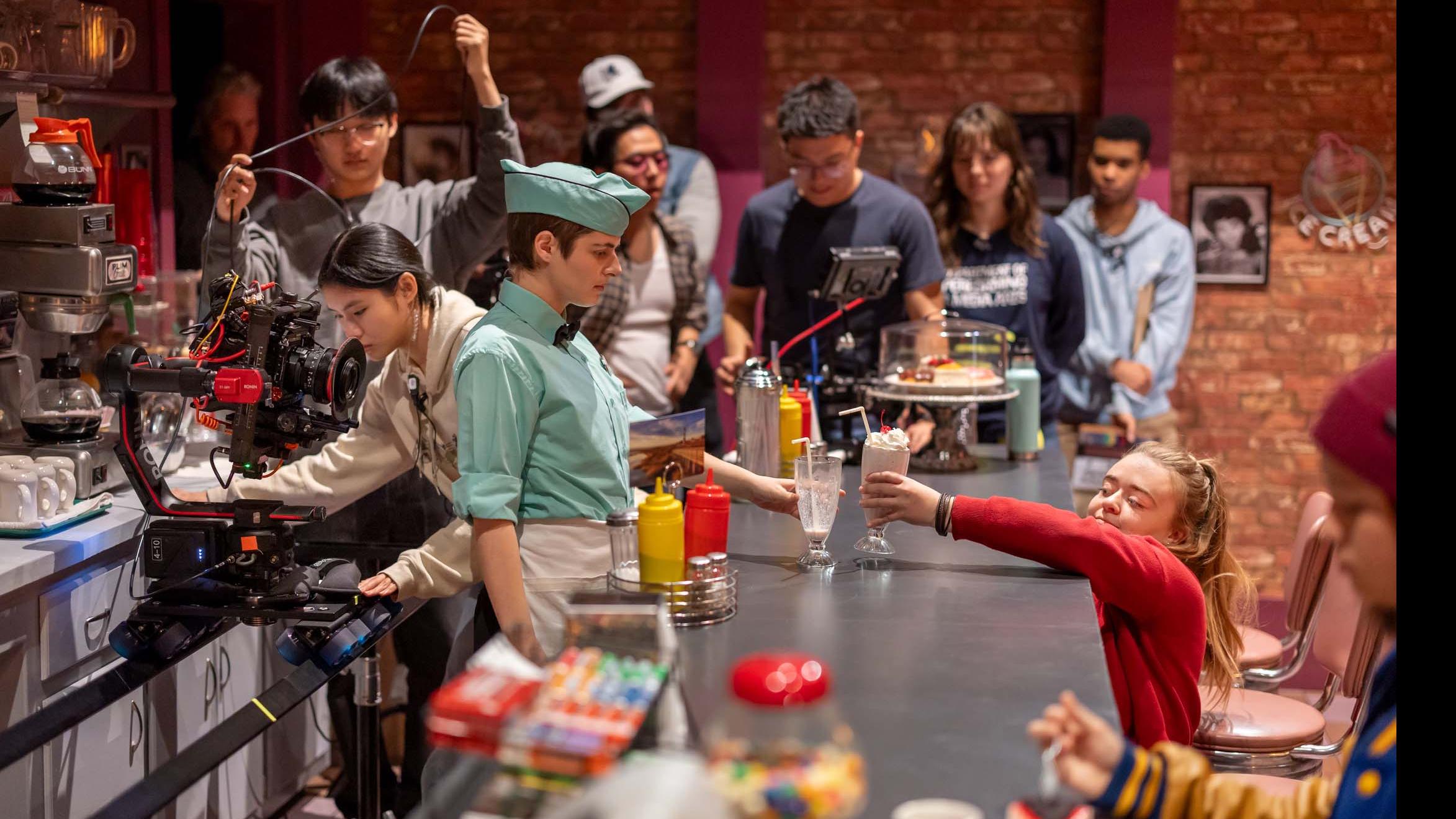 A student actor stands behind a counter while a student crew member puts a milkshake on the counter and other student crew members stand in the background on the set of the PMA Studios short film Milkshake Kiss. Photo credit: Simon Wheeler