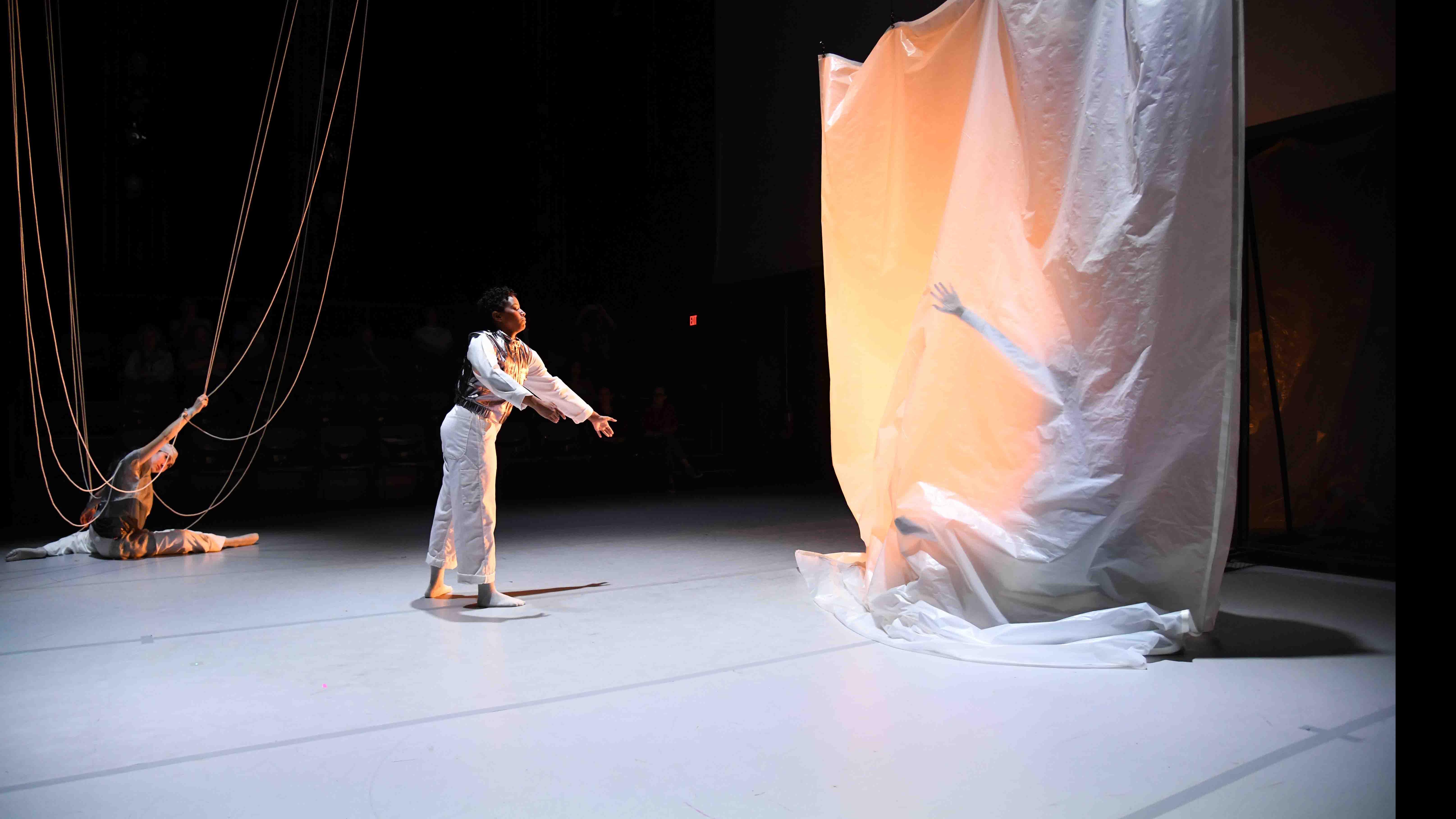 In the center, a student dancer holds their arms out towards another student dancer on the right who is behind a translucent sheet, and a student dancer on the left does a split with ropes looping down onto the stage. Photo credit: Thomas Hoebbel